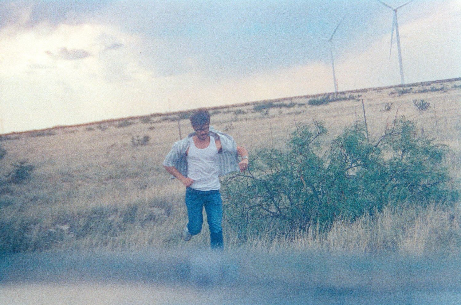 A man stands in a dry grassland field with wind turbines and cloudy sky in the background.