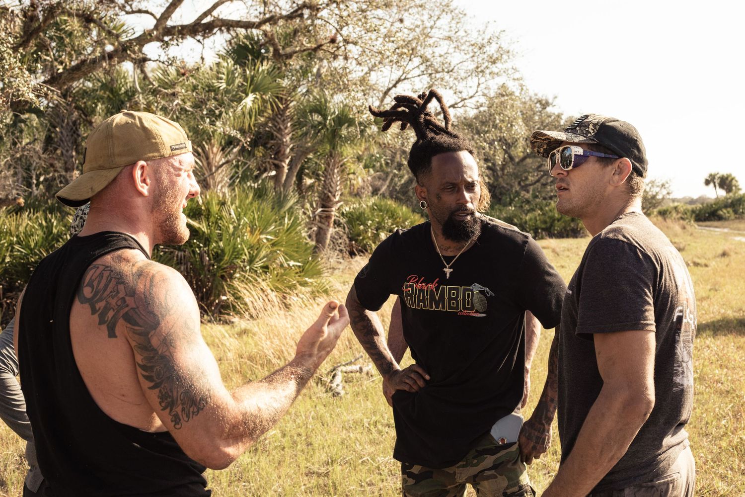 Three men having an outdoor conversation in a grassy field with trees, one with tattoos and dreadlocks.