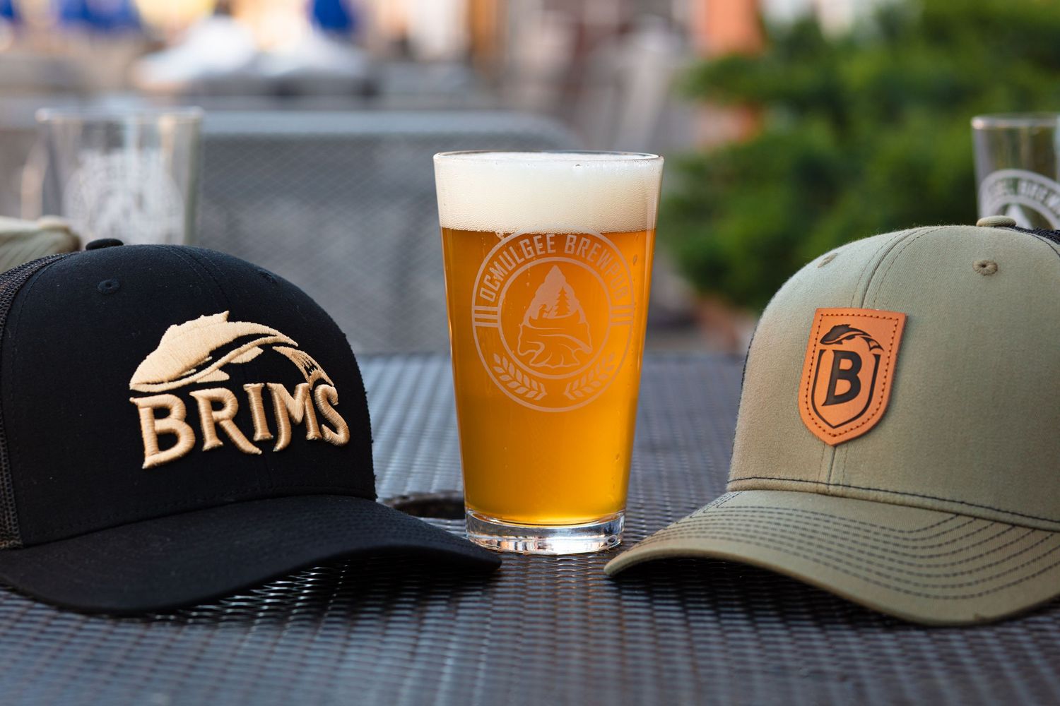 Two branded baseball caps flank a frothy craft beer glass on an outdoor patio table.