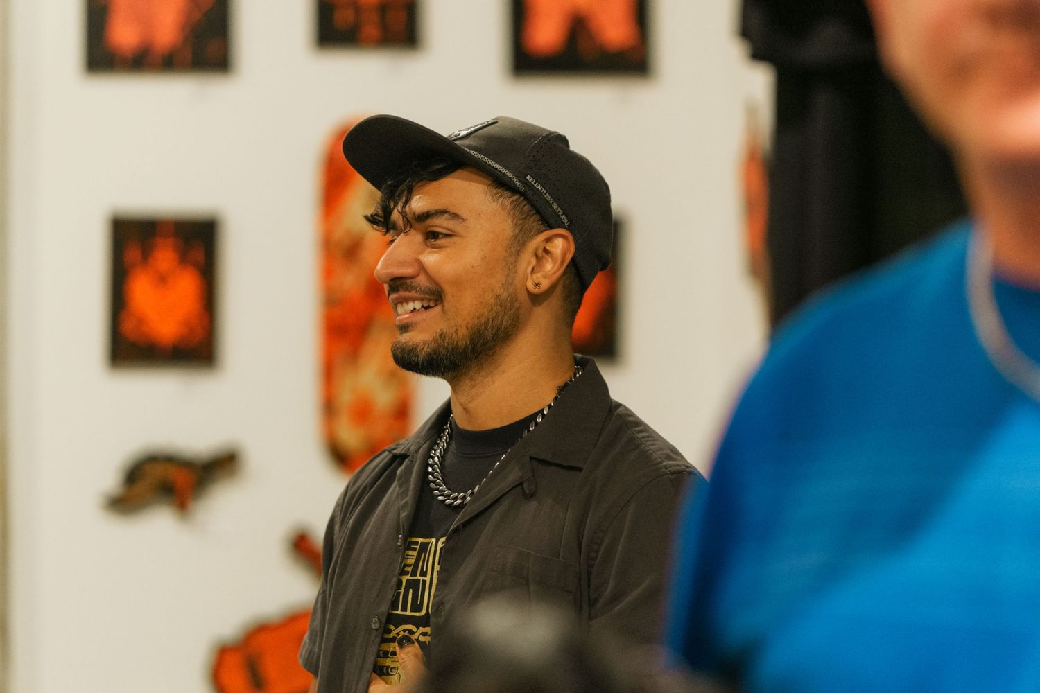 Smiling man in black cap and chain necklace stands in front of orange and black artwork.
