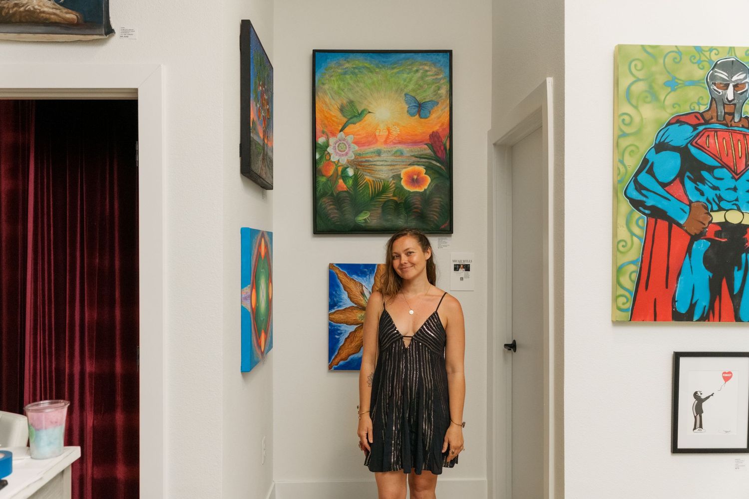 Woman in black dress stands smiling among colorful paintings in a bright art gallery.