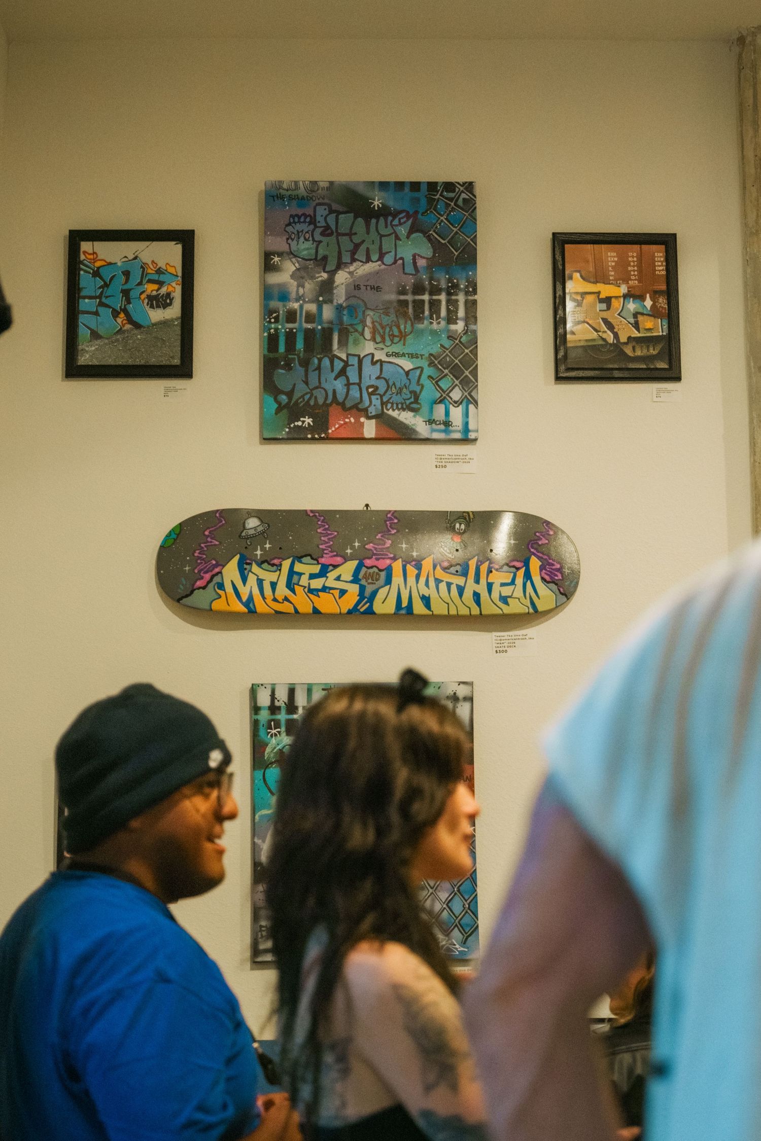 Gallery visitors admire graffiti artwork and a decorated skateboard deck reading Miles Mathew on white gallery walls.
