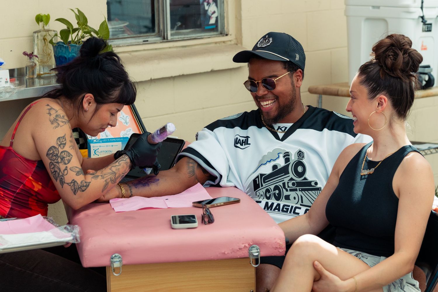 Tattoo artist carefully working on a man's arm as he smiles with a woman sitting next to him.
