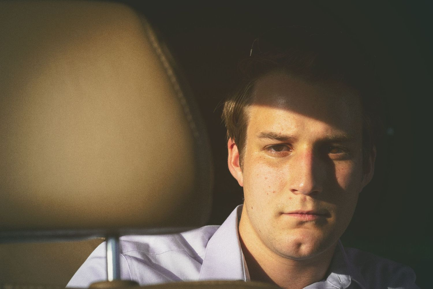 Young man in white shirt sitting in car backseat, looking serious with dramatic lighting on his face.