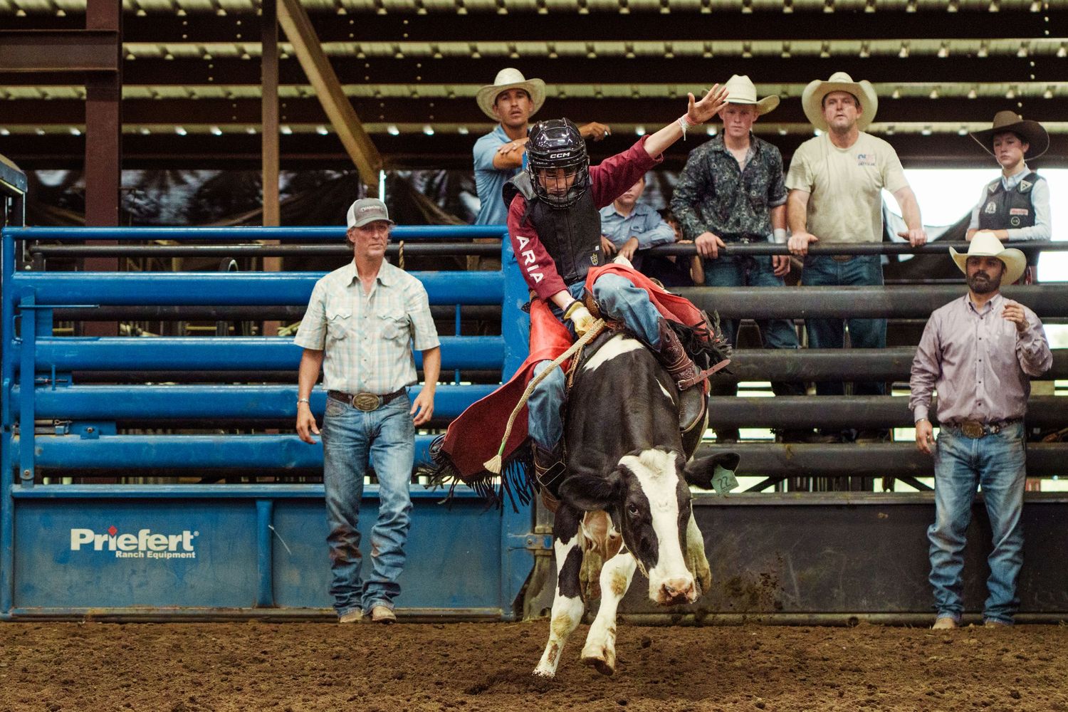 Cowboy riding a bucking bull at an indoor rodeo arena with spectators and handlers watching nearby.