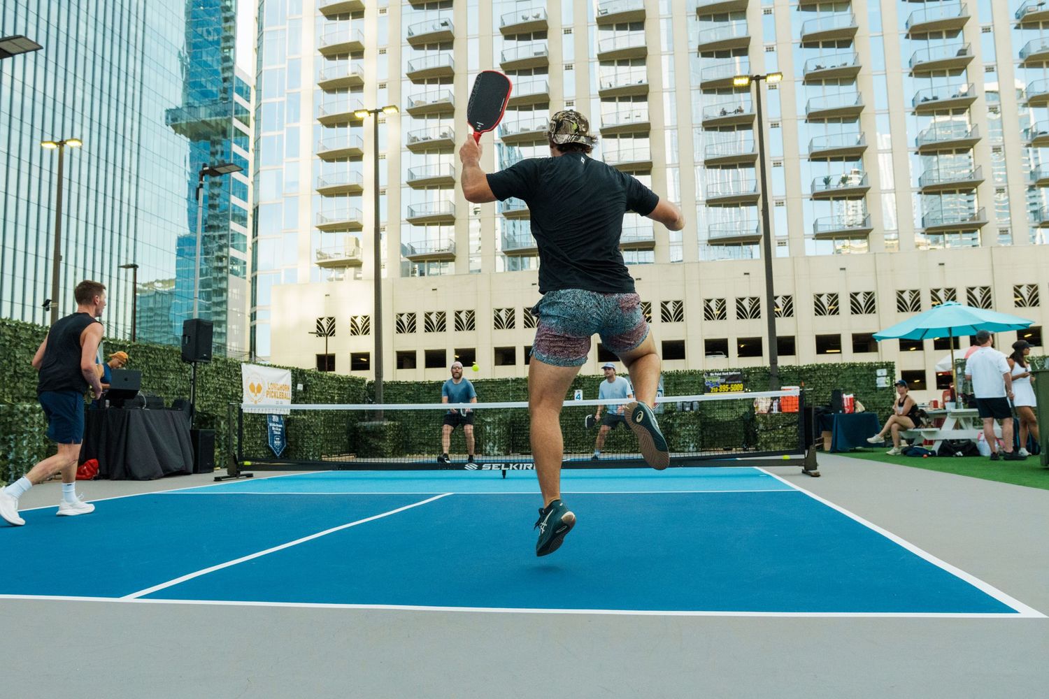 Player leaping mid-air on an outdoor pickleball court with urban skyscrapers in the background.