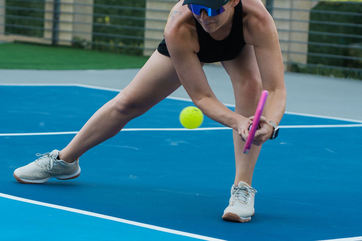 Woman playing pickleball on blue court, lunging to hit yellow ball with pink paddle while wearing blue goggles.