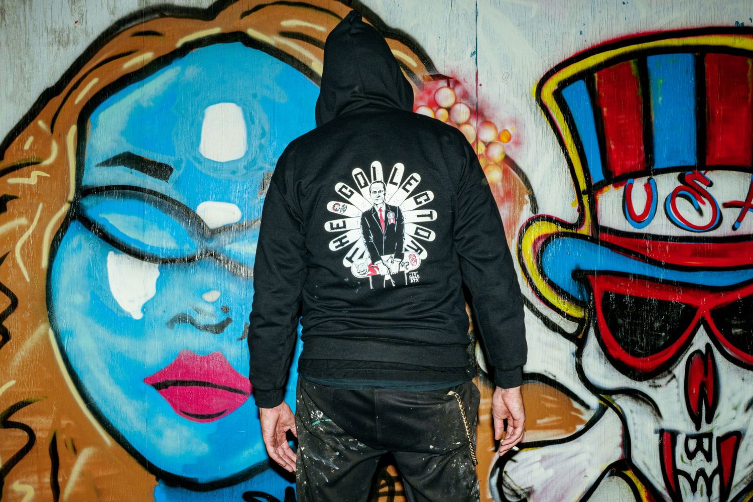 Person in black graphic hoodie standing before colorful graffiti mural with blue face and skull art.