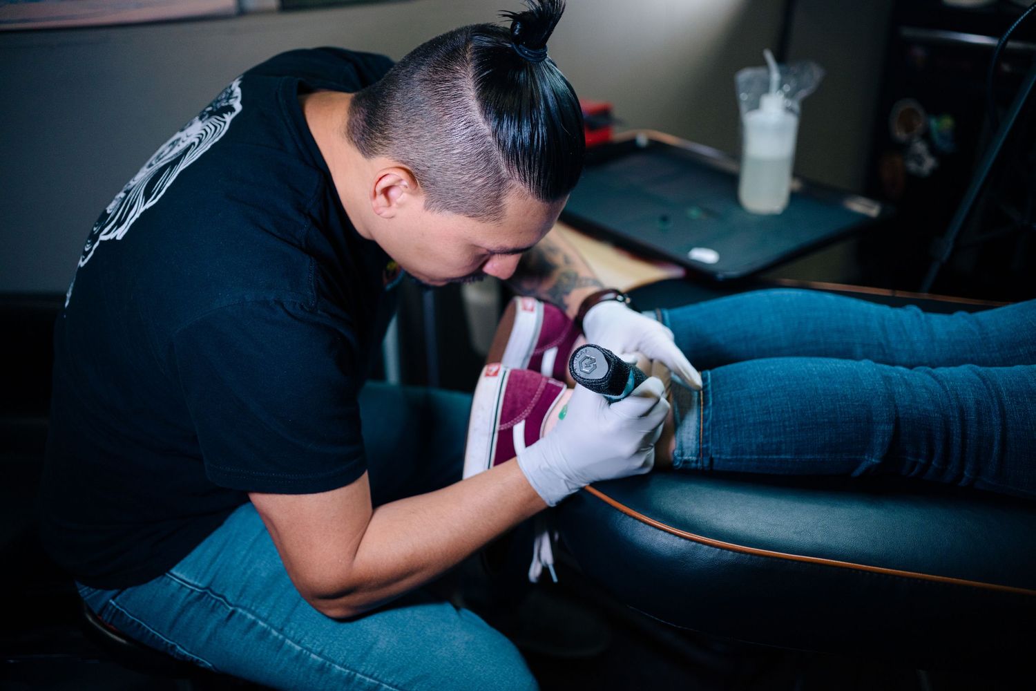 A tattoo artist carefully works on a client's leg in a professional tattoo studio, wearing gloves and holding a tattoo machine.