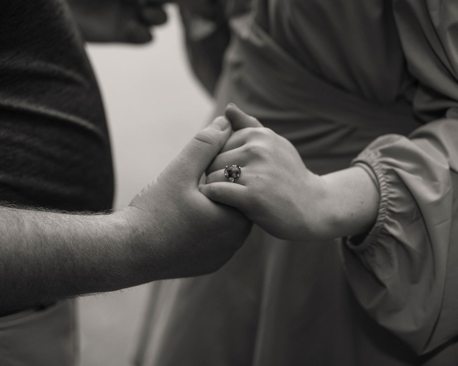 Close-up black and white photo of a couple holding hands, woman wearing a diamond engagement ring.