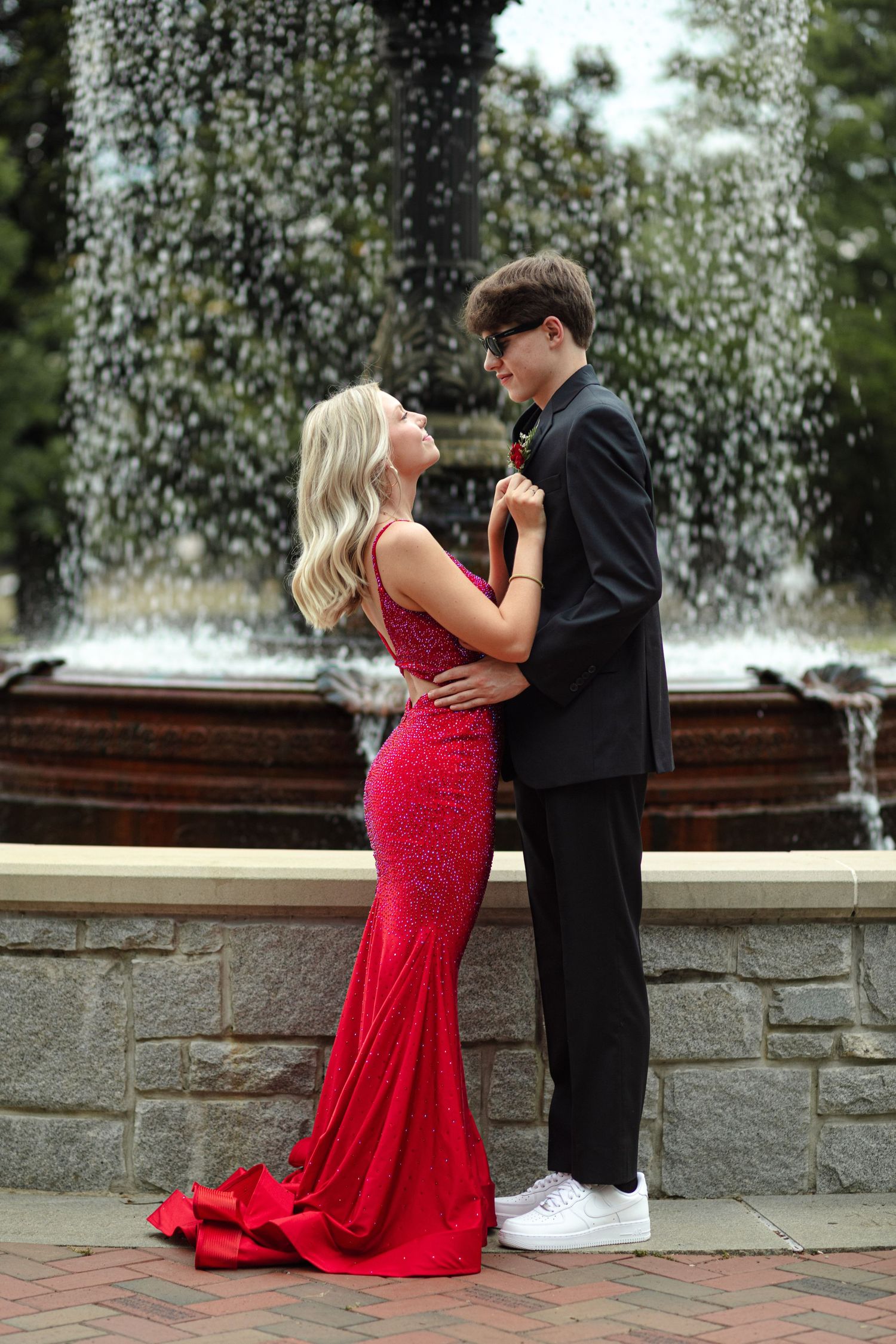Couple in prom attire posing by a fountain, girl in red sequin gown and boy in black suit.