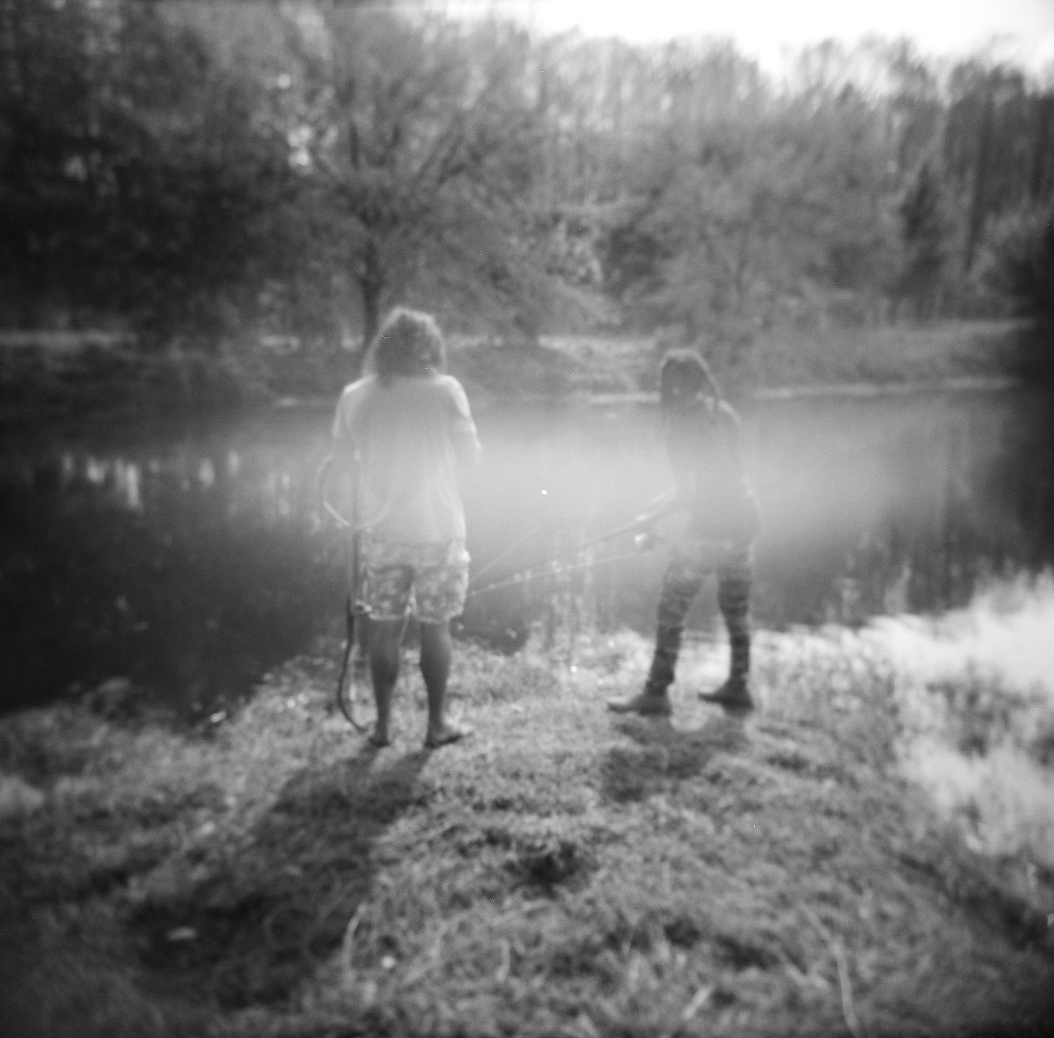 Two children stand by a reflective pond surrounded by trees in a dreamy black and white photo.