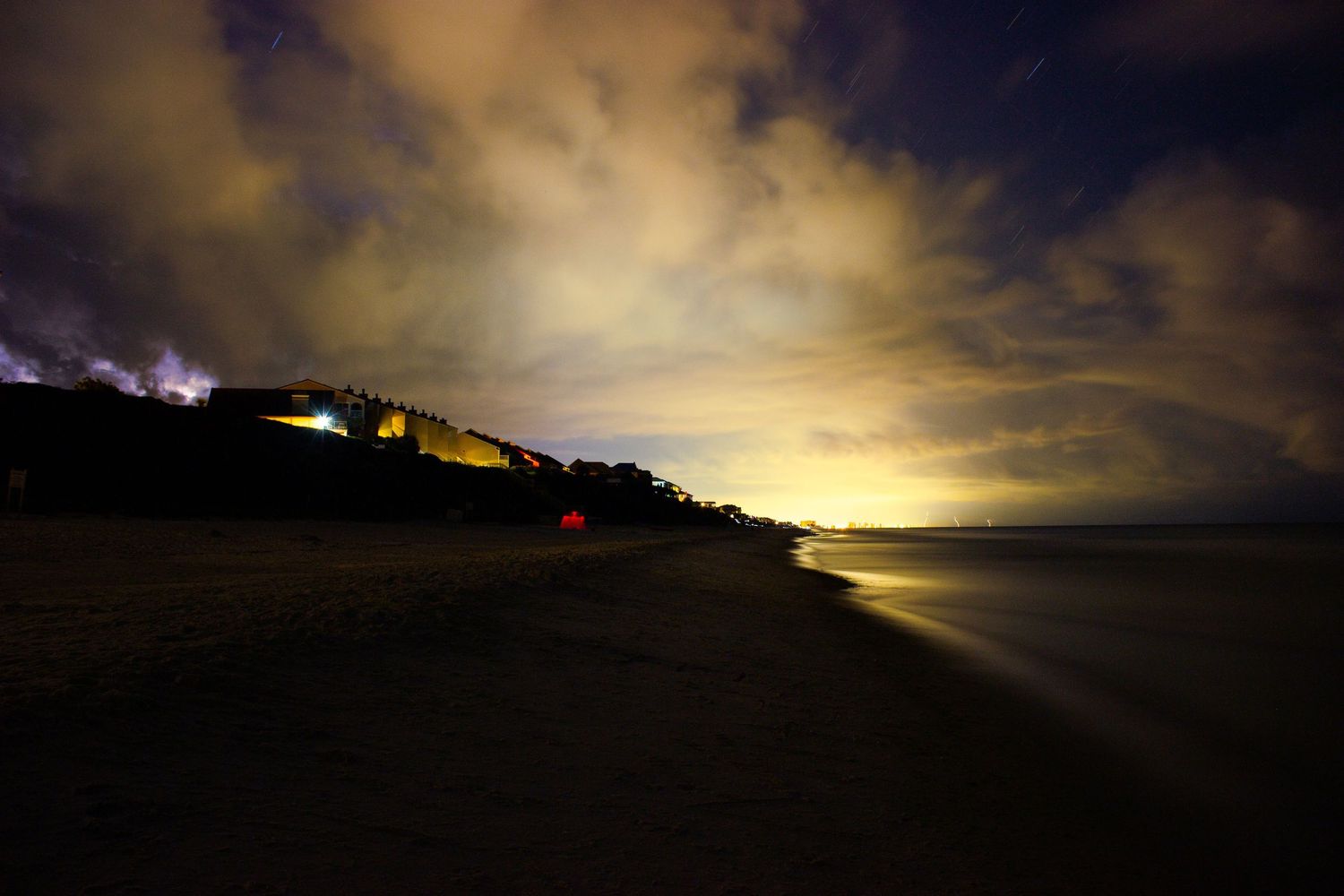 Nighttime beach scene with dark sandy shore, calm ocean waters, and glowing city lights illuminating cloudy sky.