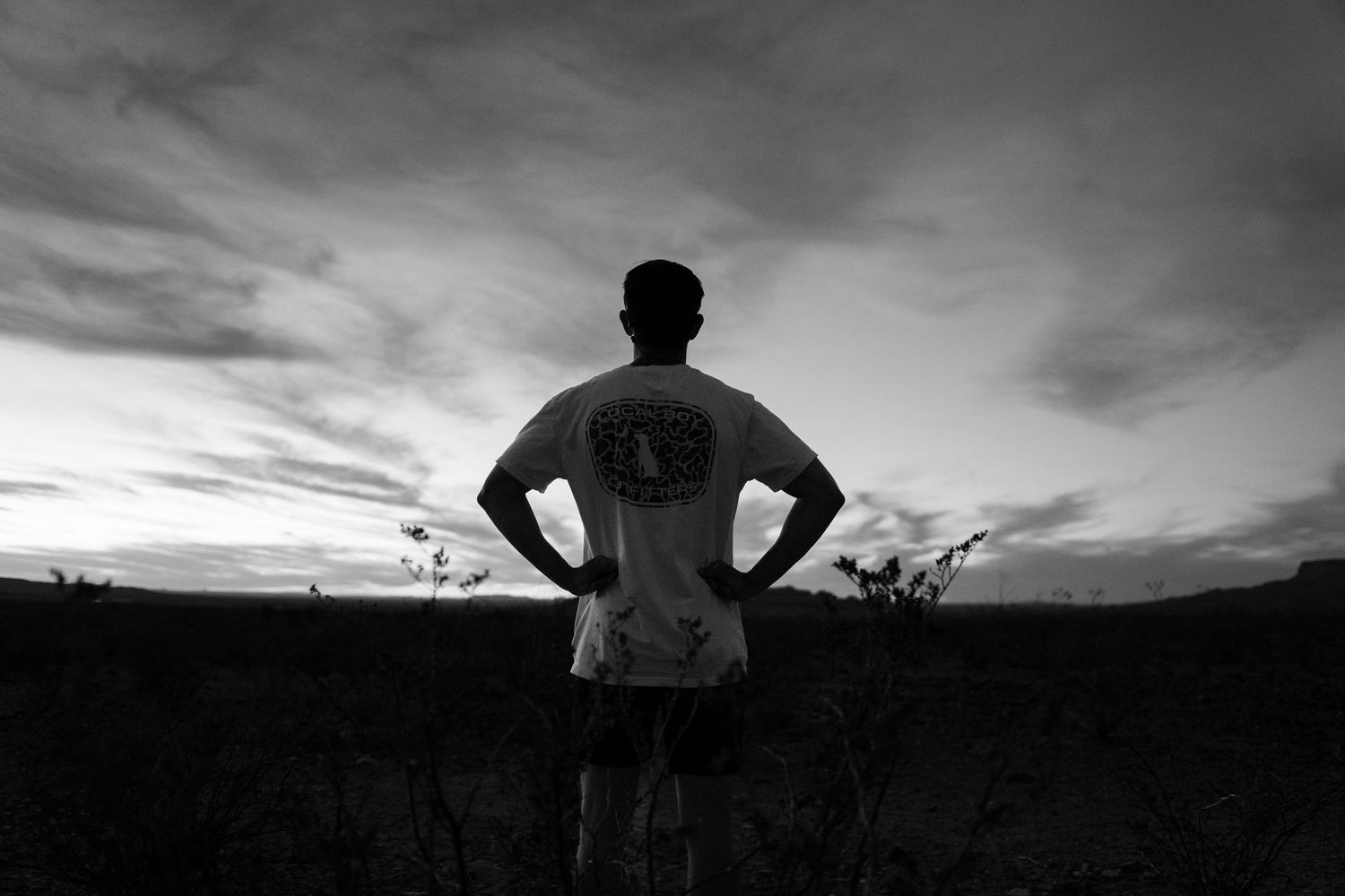 Black and white photo of a man standing in a field with hands on hips, silhouetted against a dramatic cloudy sky.