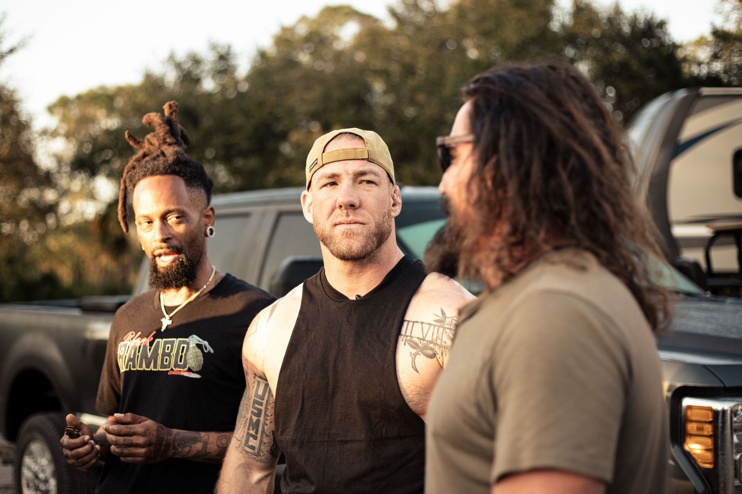 Three tattooed men with dreadlocks and casual attire conversing outdoors near a truck at golden hour.