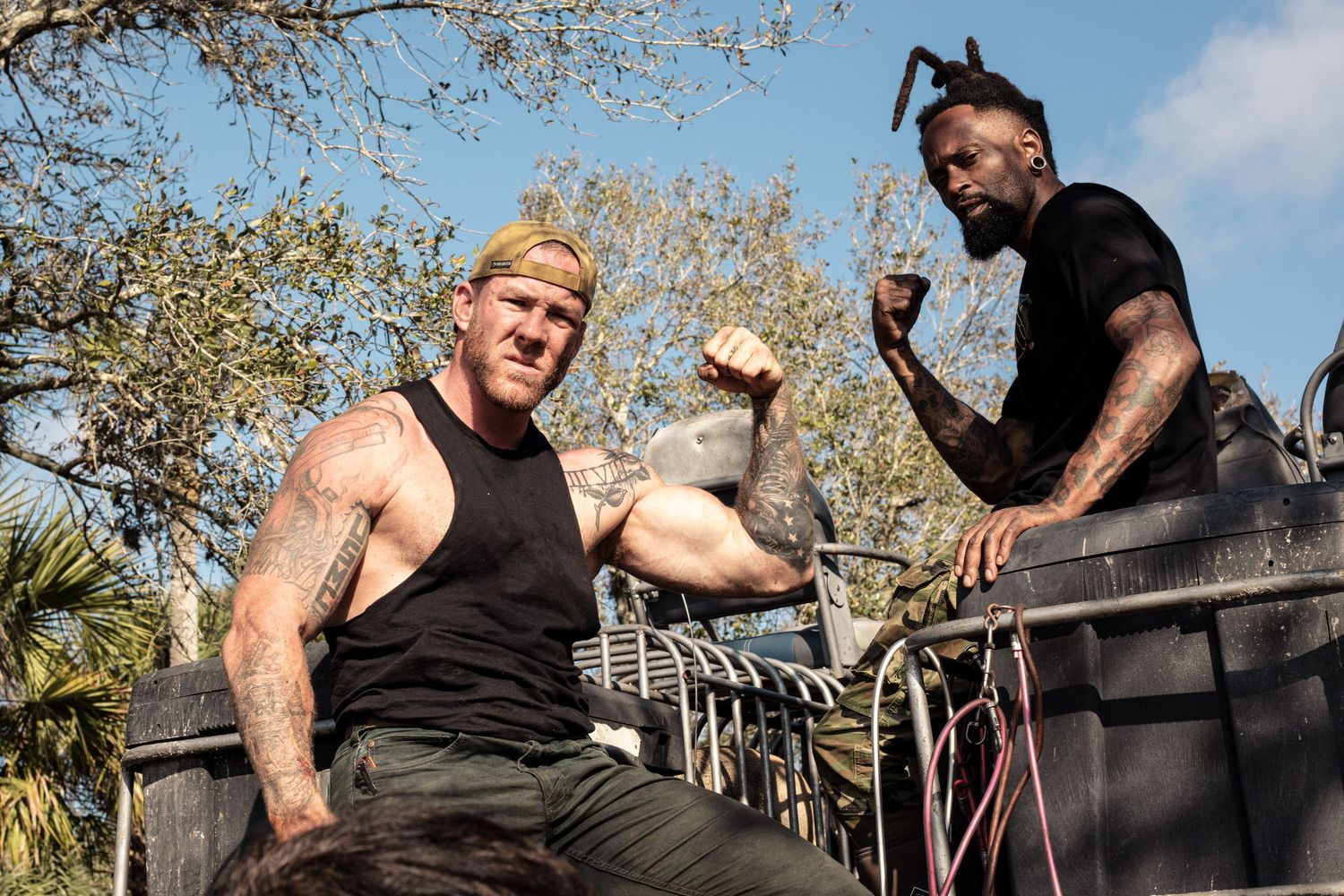Two tattooed muscular men flexing on the back of a truck outdoors on a sunny day.