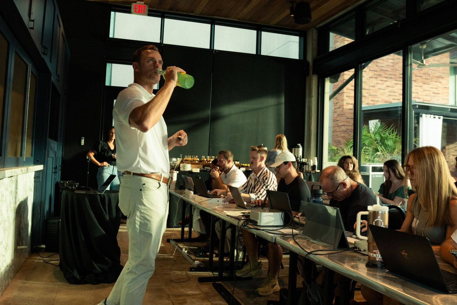 Man in white outfit speaks to guests seated at long tables in a modern restaurant venue.