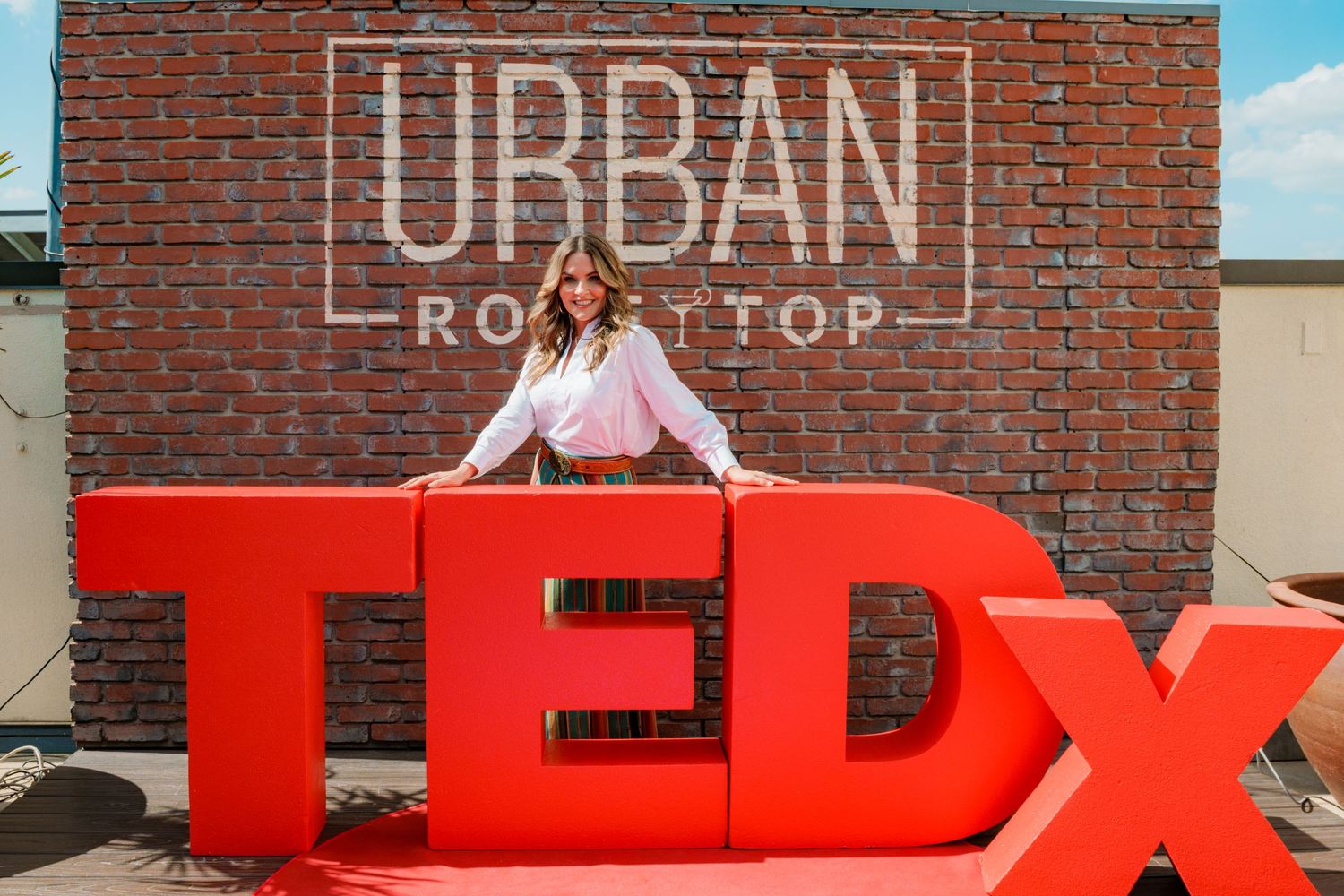 Woman posing at TEDx event on Urban Rooftop with large red TEDx letters and brick wall backdrop.