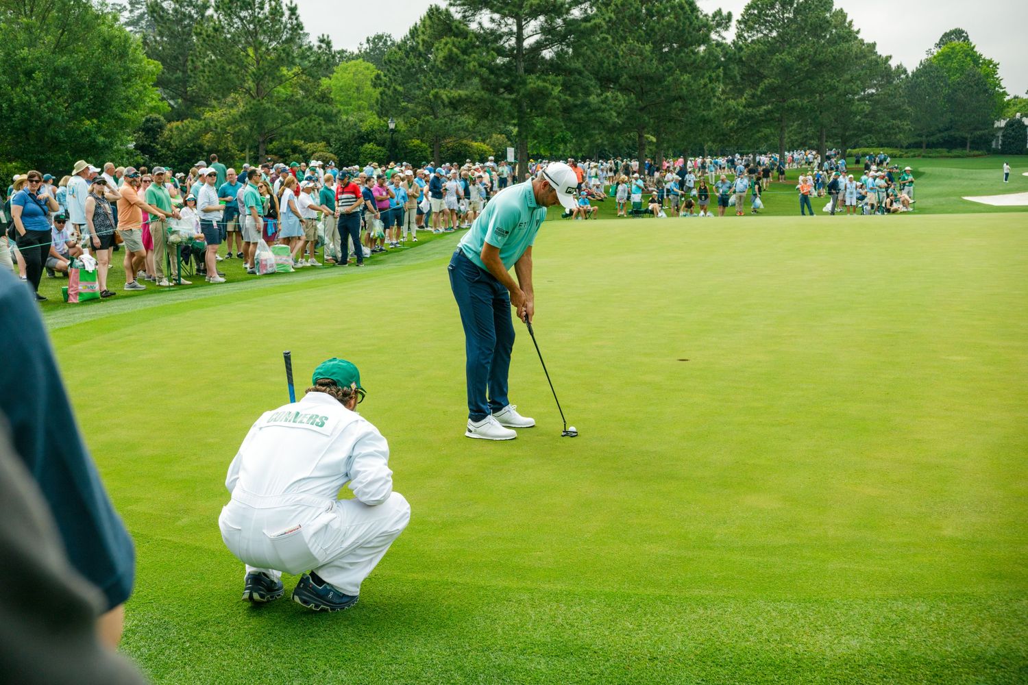 Golfer putting on green at Masters Tournament with caddie crouching nearby, surrounded by large gallery of spectators.