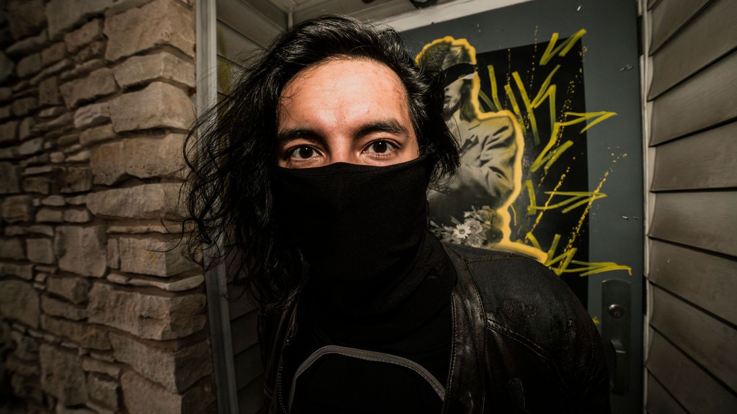 Person with long dark hair wearing black face mask standing near graffiti wall.
