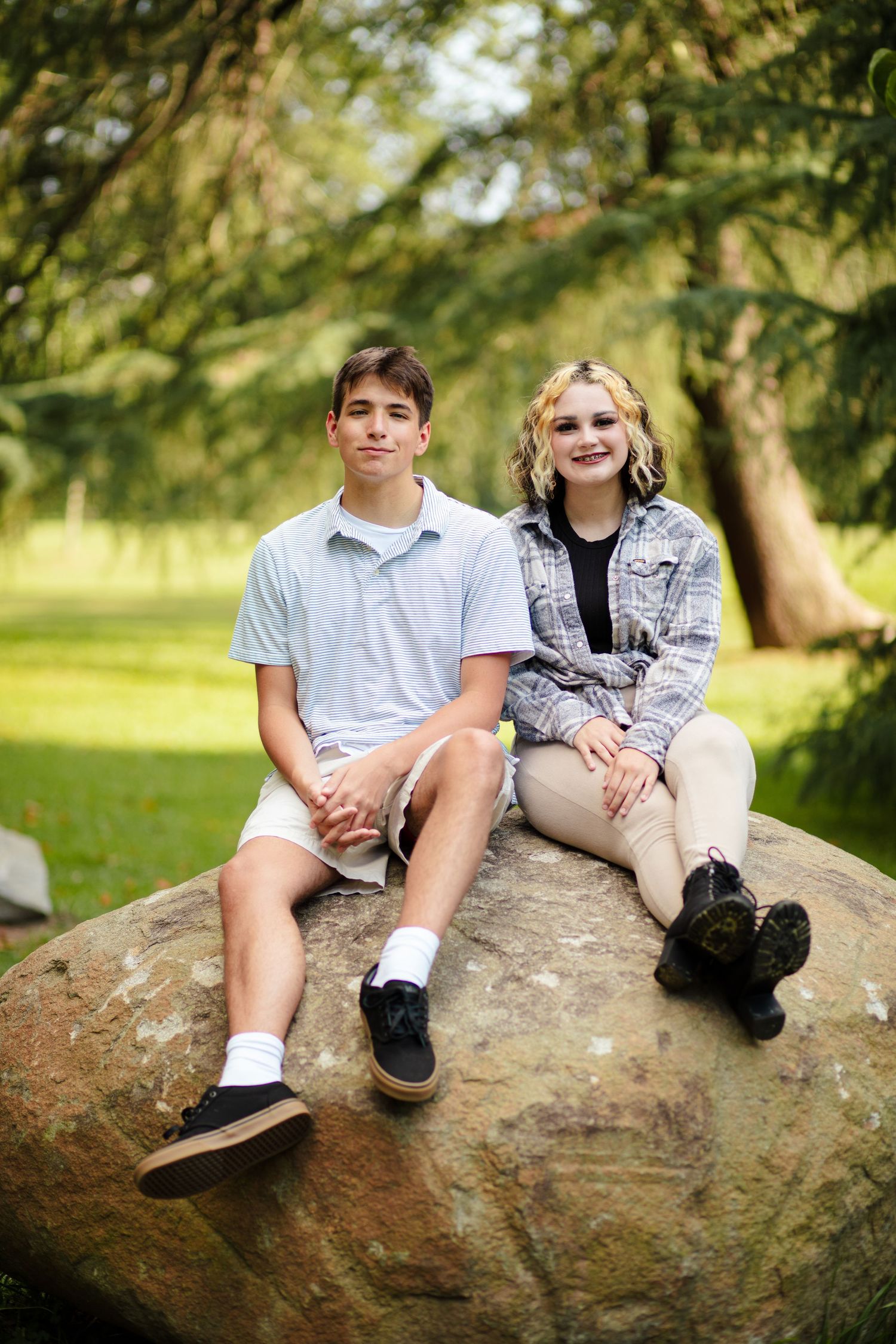 Teen boy and girl sitting together on large rock in lush green park.