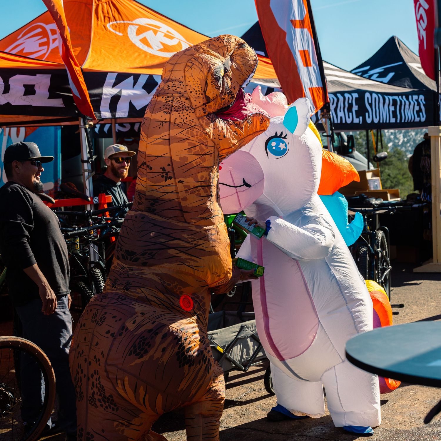 Two people in inflatable T-Rex and unicorn costumes at an outdoor mountain bike festival.