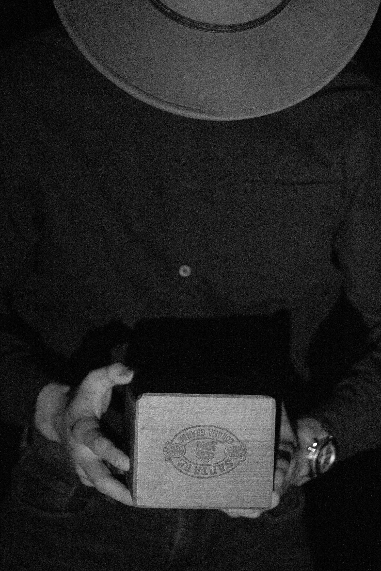 Black and white photo of a person in a cowboy hat holding a vintage cigar box, wearing a watch.