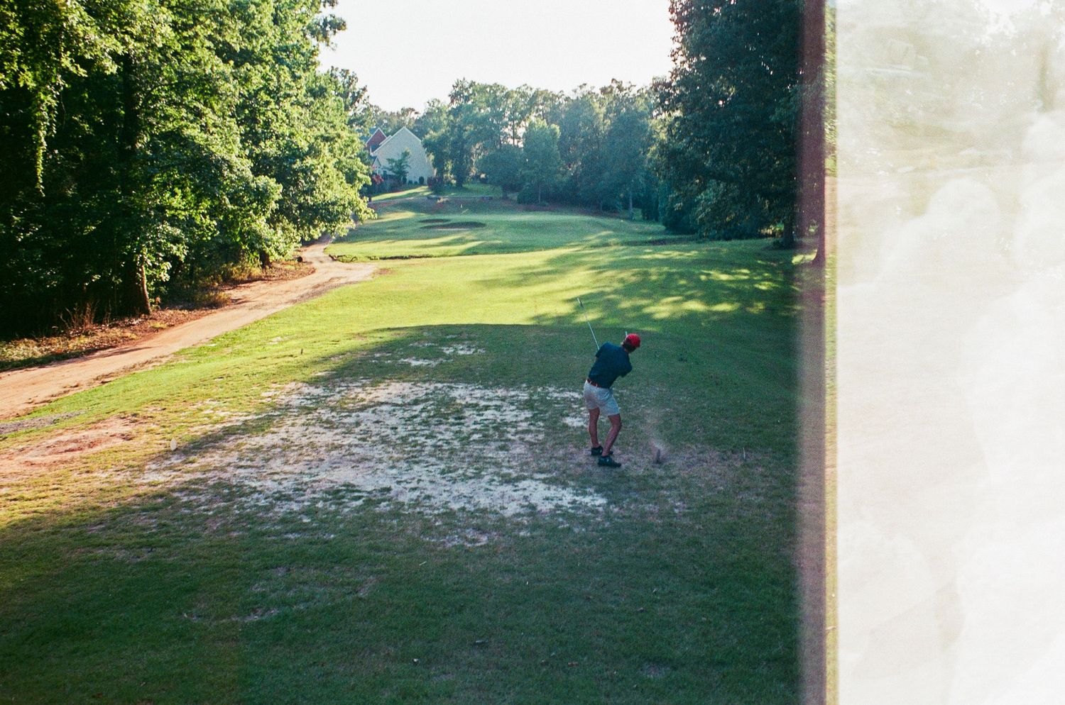 Golfer hitting a shot from a sandy area on a scenic tree-lined golf course fairway.