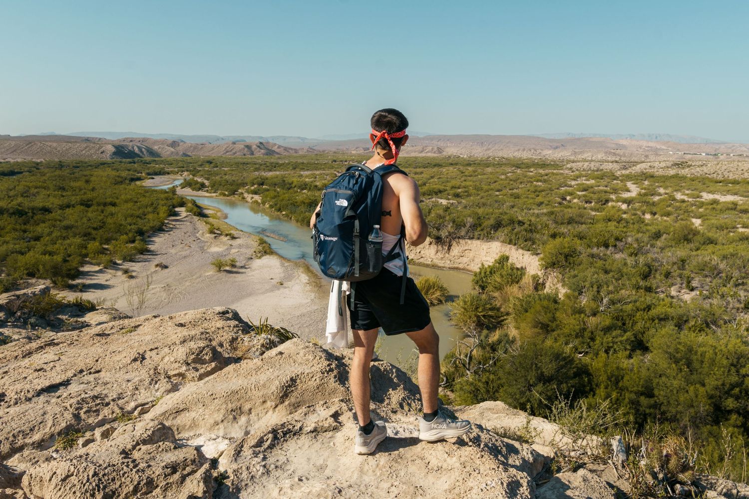 Hiker with blue backpack stands on rocky overlook surveying a river valley and vast desert landscape.
