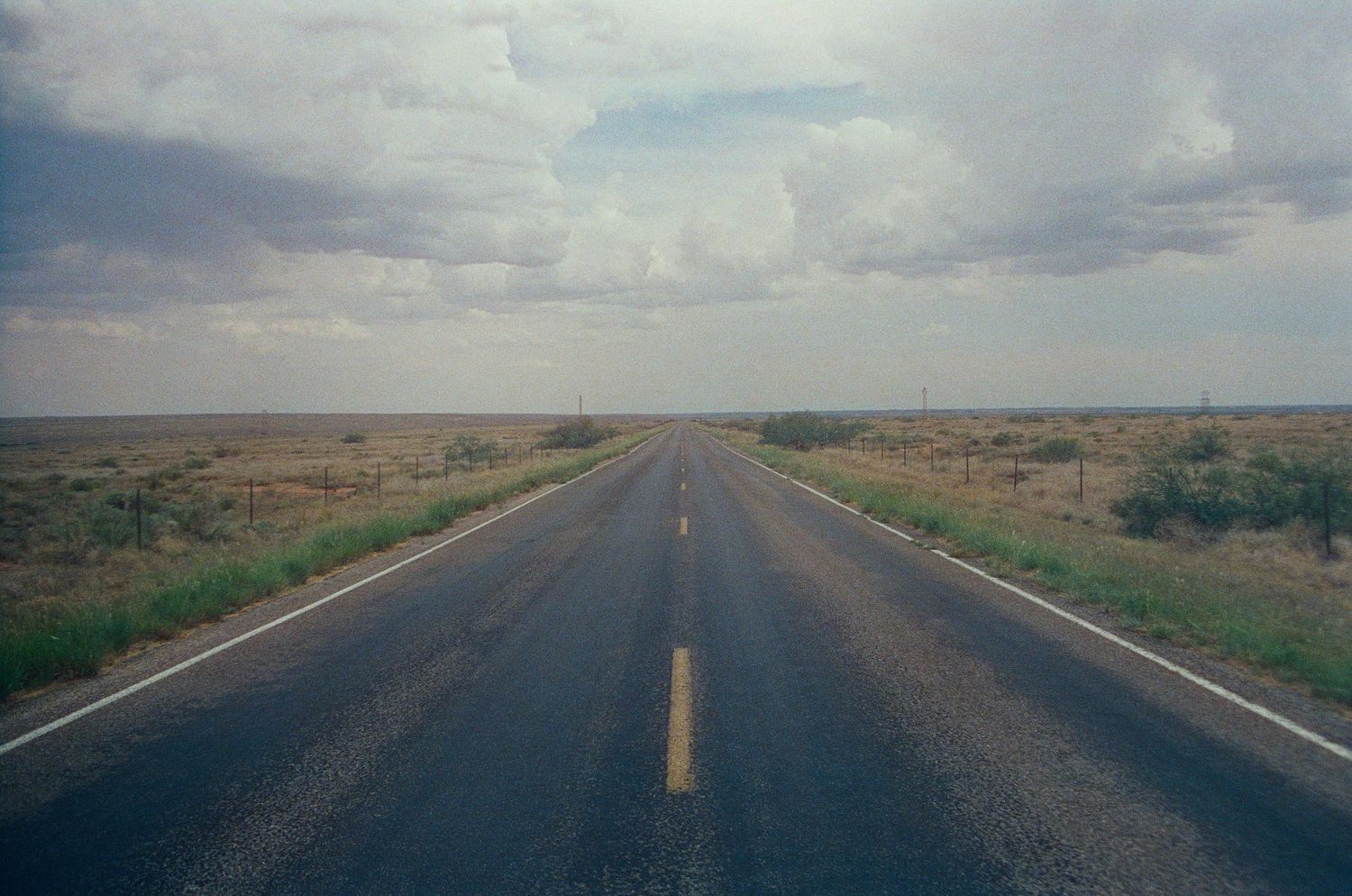 A straight two-lane highway stretches to the horizon through flat arid plains under a dramatic cloudy sky.