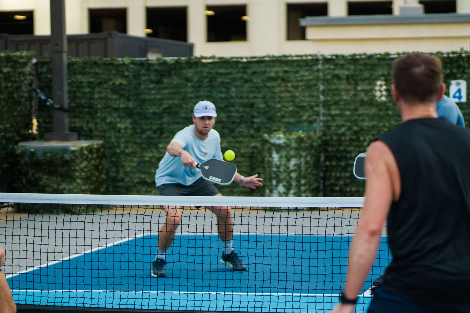 Two men playing pickleball on a blue outdoor court, one preparing to hit a yellow ball near the net.