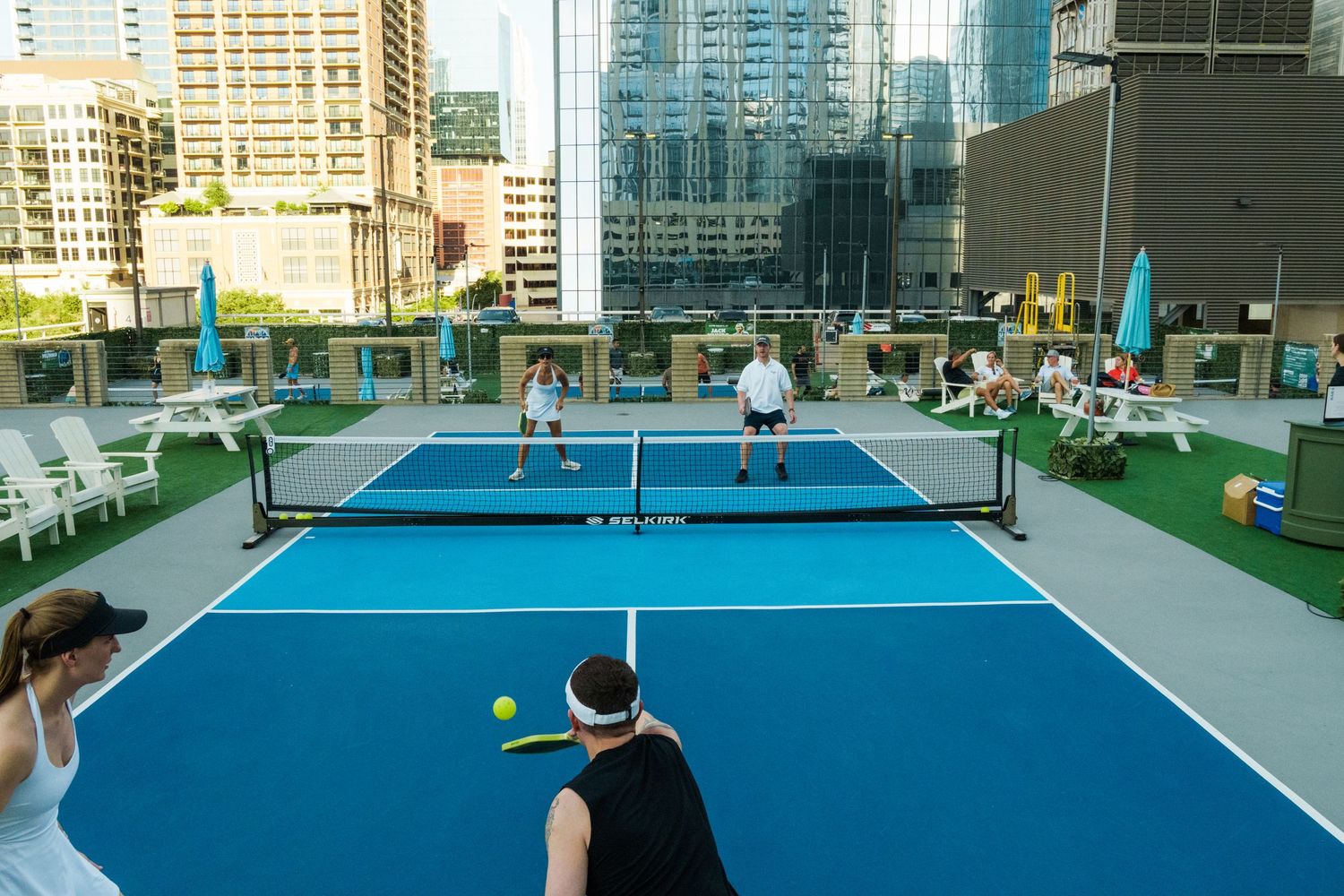 Players enjoy a rooftop pickleball court with blue surface, net, and city skyline backdrop on a sunny day.