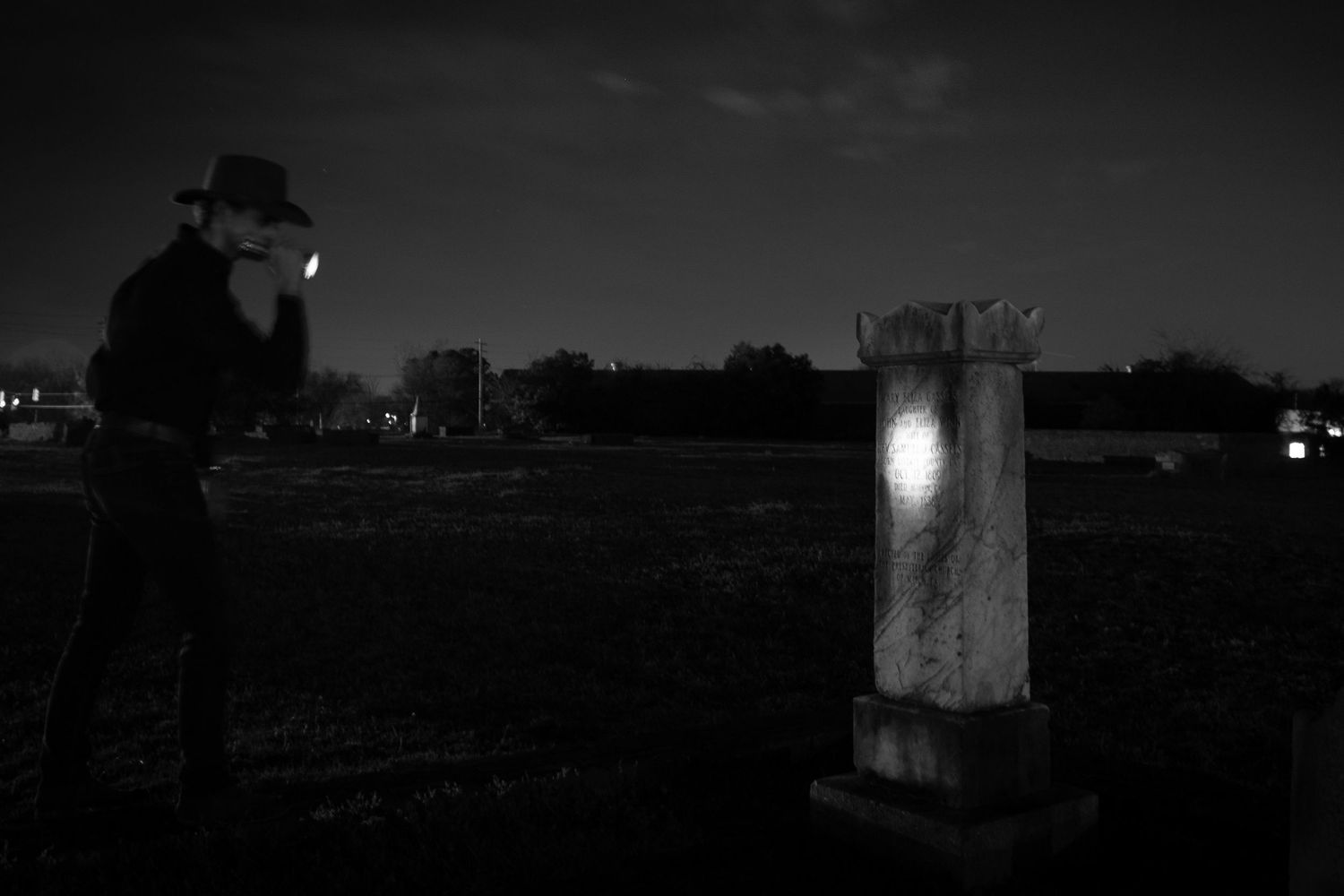 Man in hat using phone flashlight to examine gravestone in dark cemetery at night.
