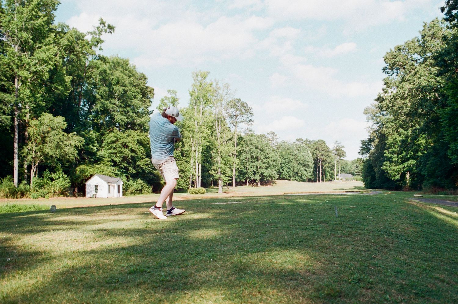 Golfer swinging a club on a lush green golf course surrounded by tall trees on a sunny day.