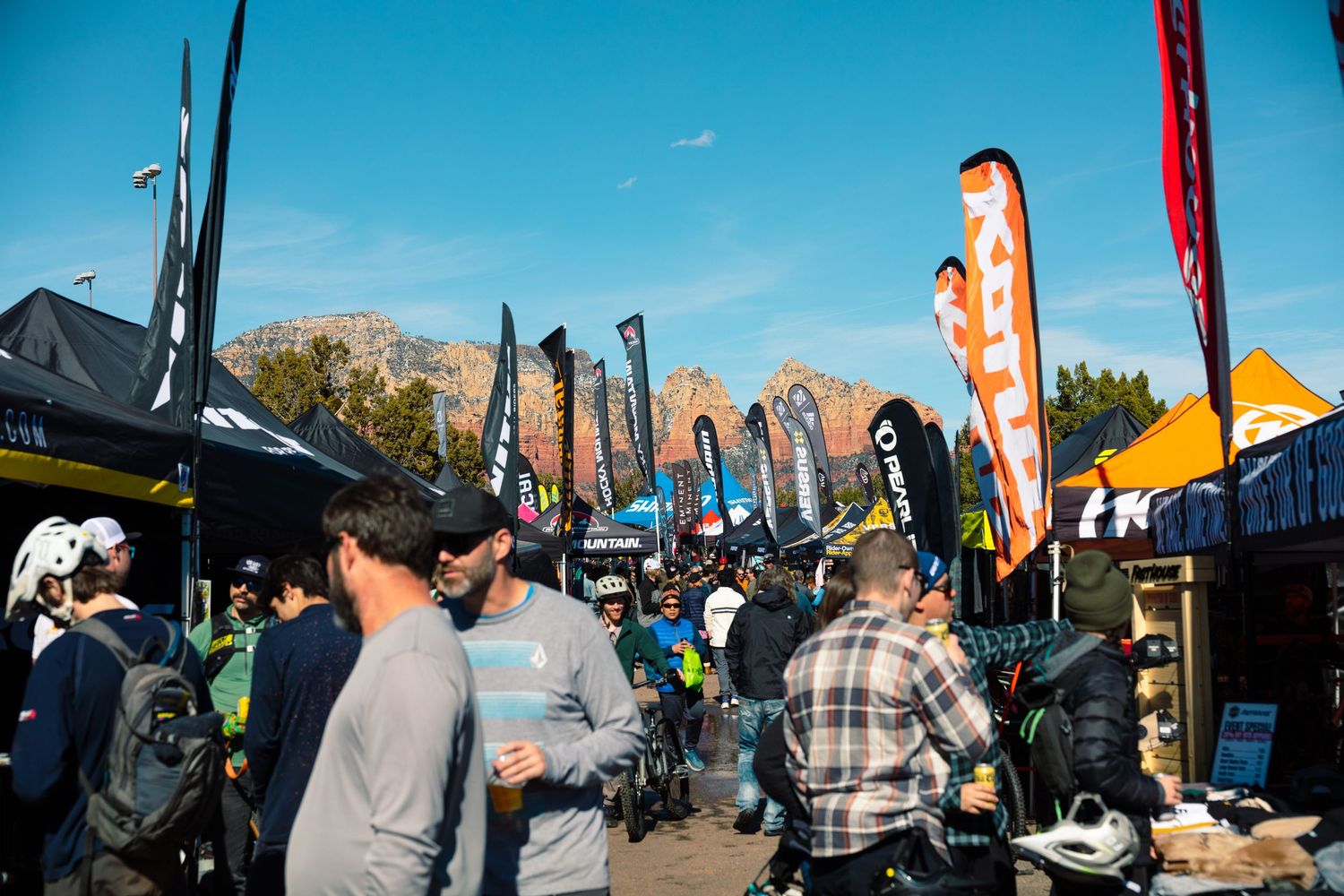 Large crowd browses vendor booths with tall banner flags at an outdoor mountain bike expo event.