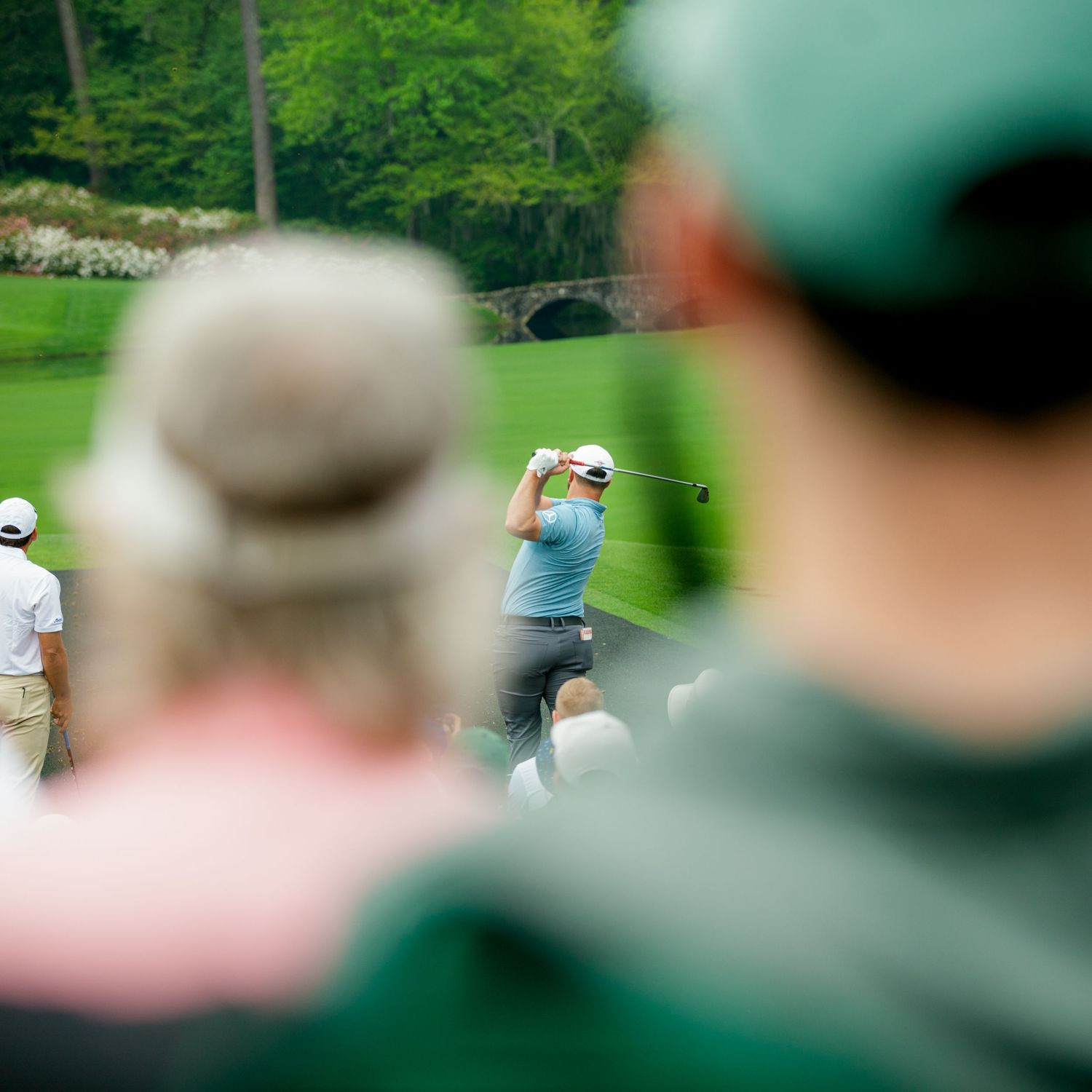 Spectators watch a golfer swing at a lush golf course during a tournament, with a stone bridge visible in the background.