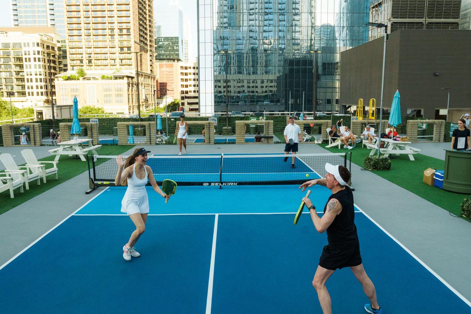 Players enjoy a rooftop pickleball court with stunning city skyline views on a bright sunny day.