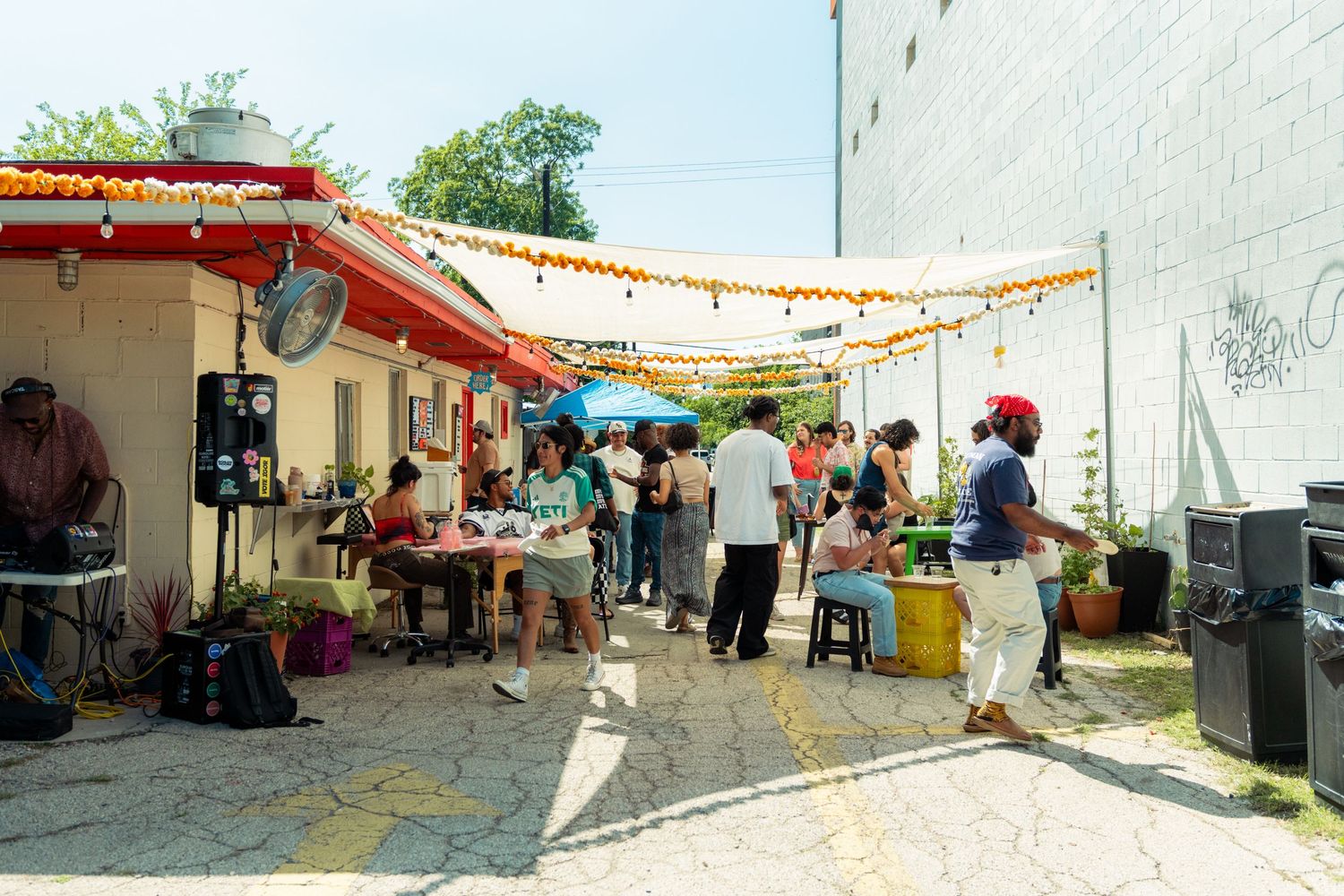 Lively outdoor community event with people gathered near a food stand decorated with string lights.