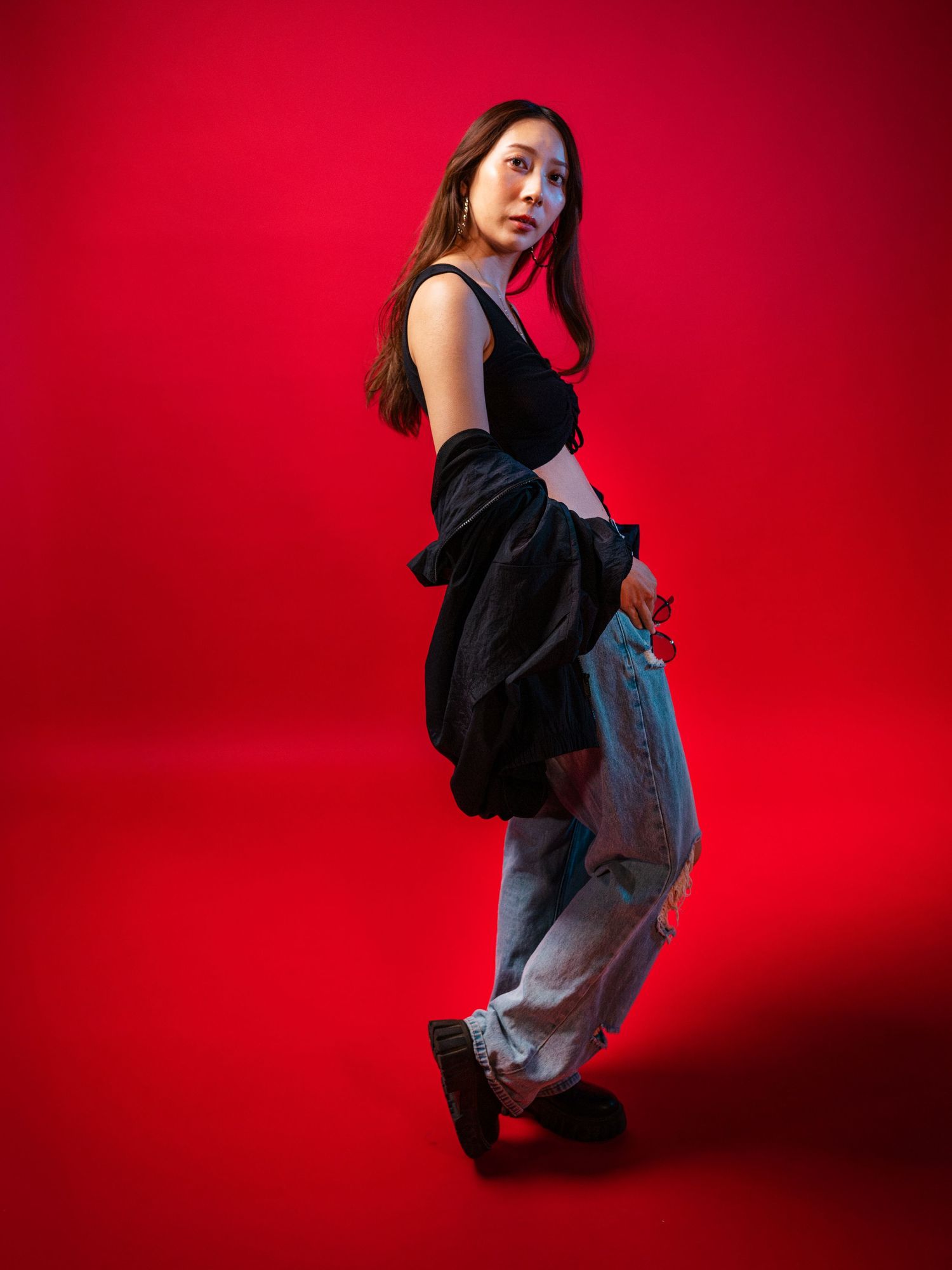 Asian woman in black crop top and ripped jeans poses sideways against a striking red studio background.