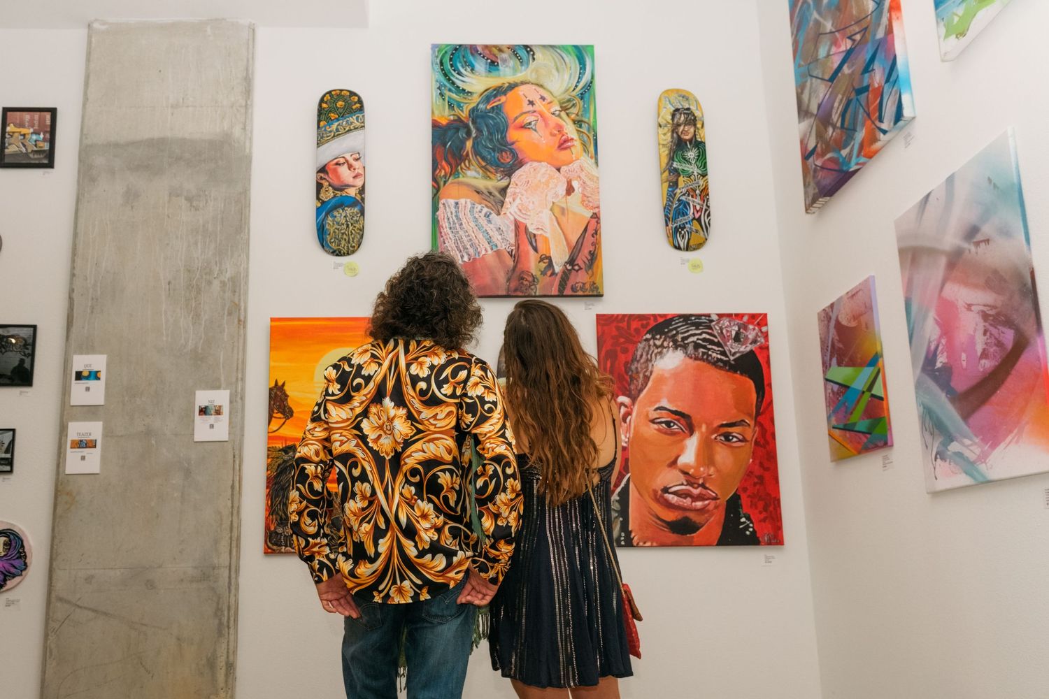 Two visitors viewing large colorful portrait paintings at an art gallery exhibition.