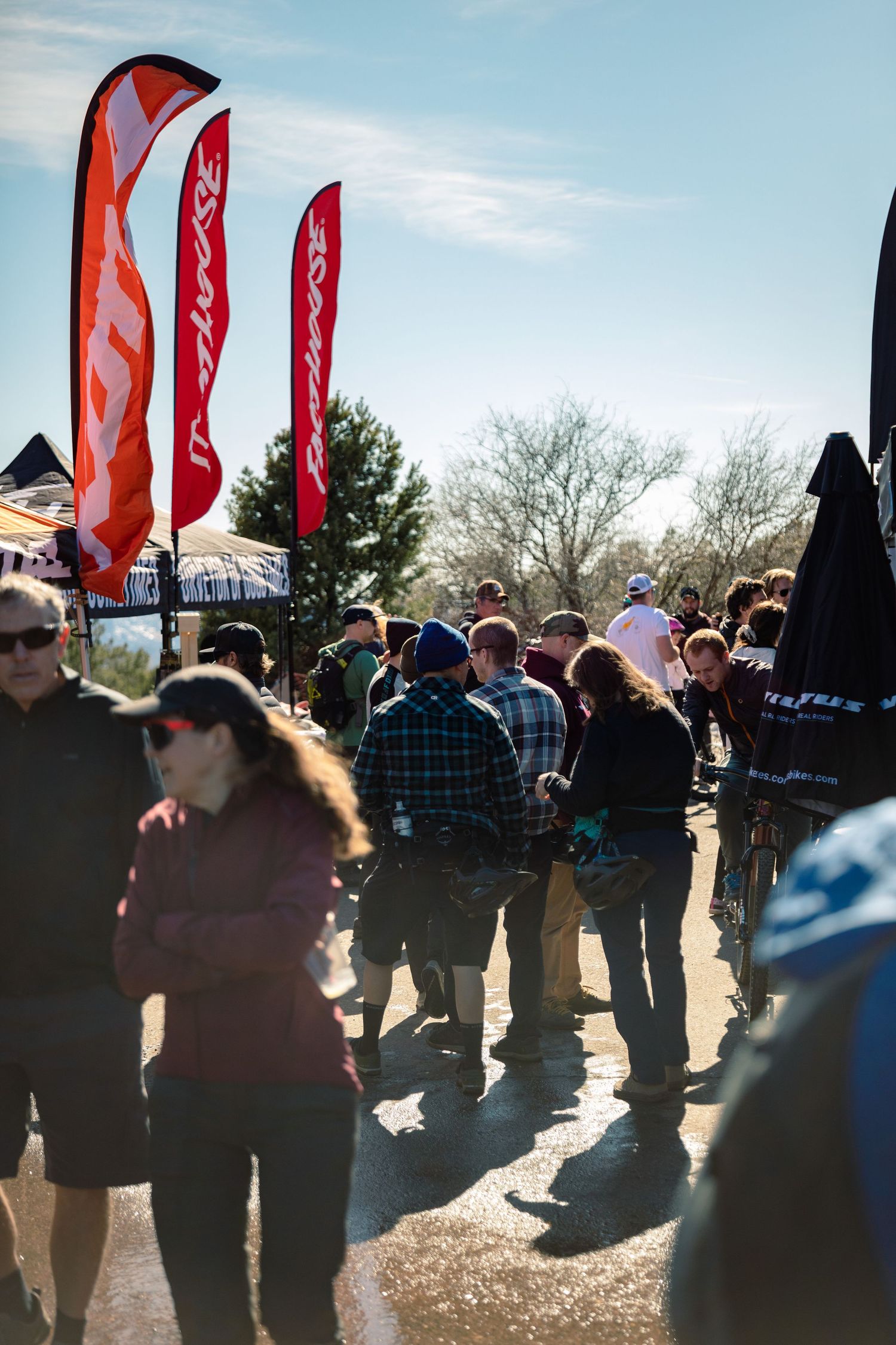 Crowd of cycling enthusiasts walks among vendor booths with colorful banner flags at an outdoor expo.