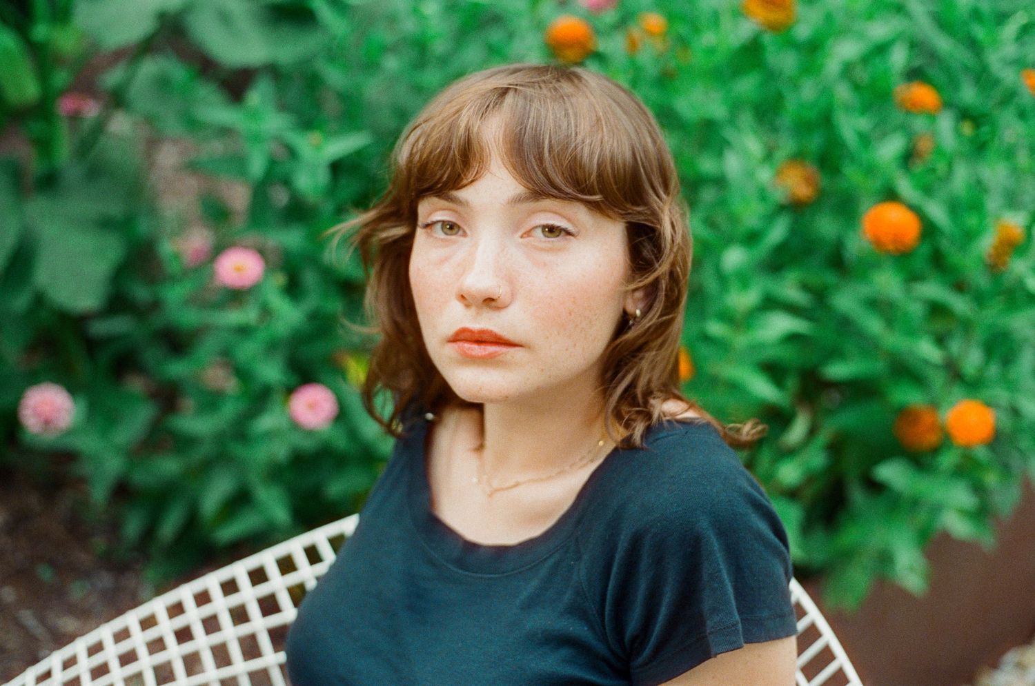 Young woman with short brown hair and bangs sits outdoors among vibrant green foliage and orange flowers.