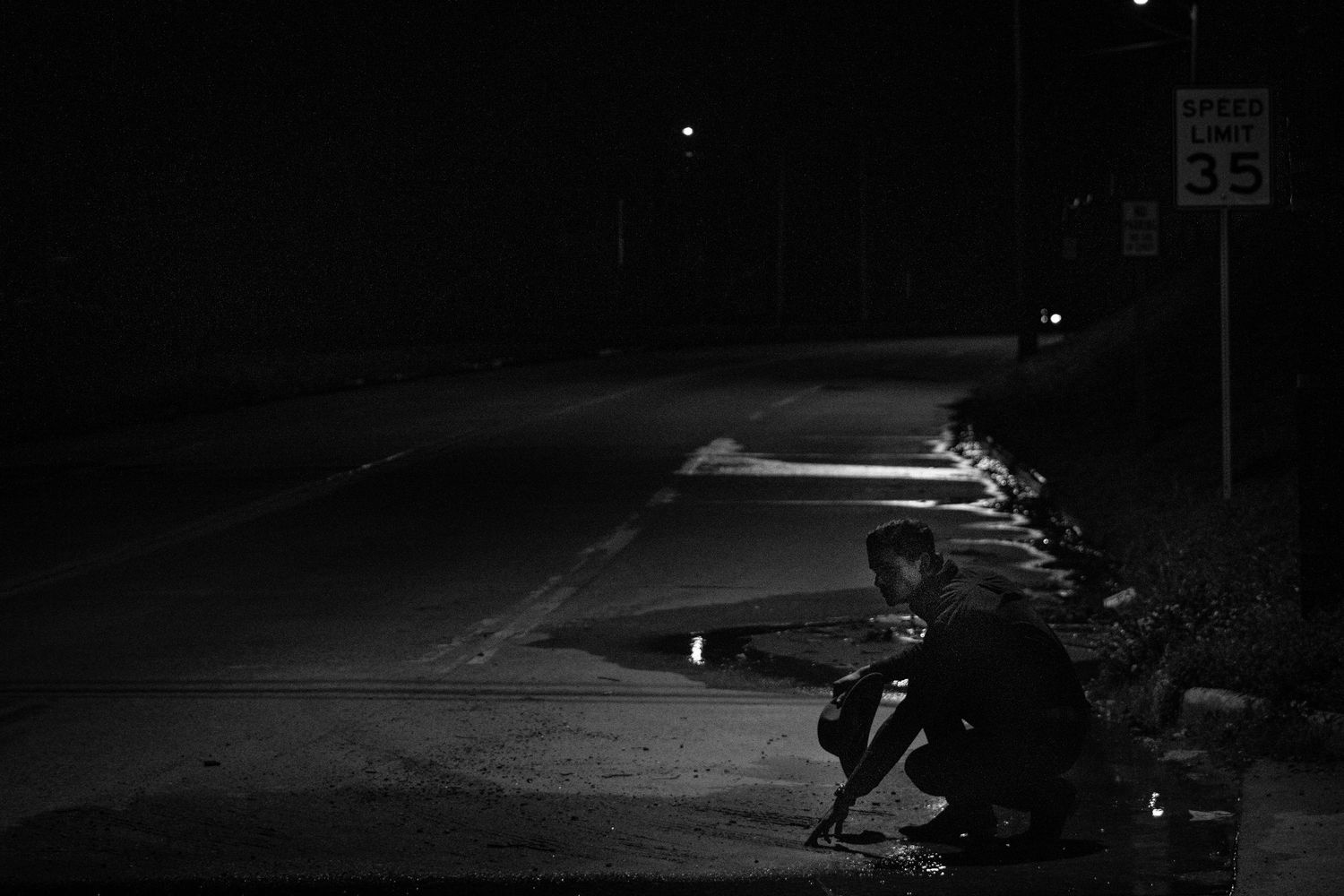 Person crouching by roadside at night near speed limit sign in black and white.