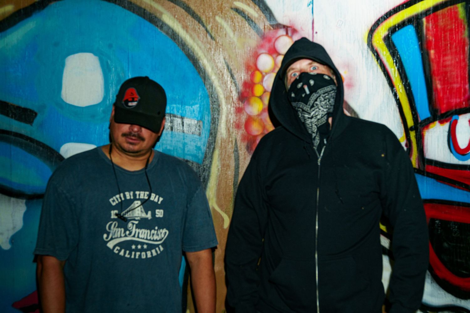 Two men posing in front of colorful graffiti wall, one wearing a bandana mask and hoodie in urban setting.