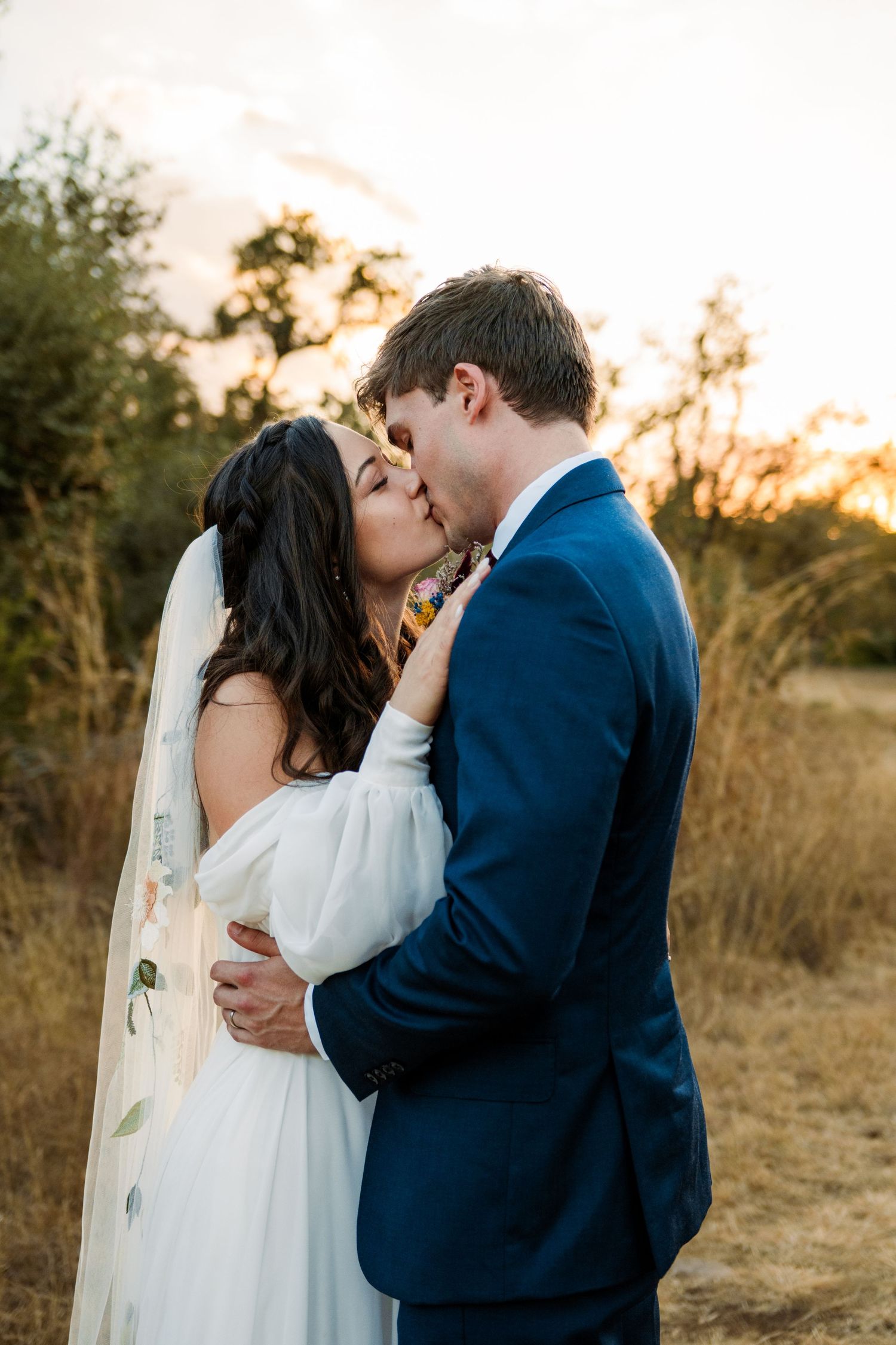 Bride and groom sharing a romantic kiss at sunset in a golden field, bride in white gown with veil, groom in navy suit.
