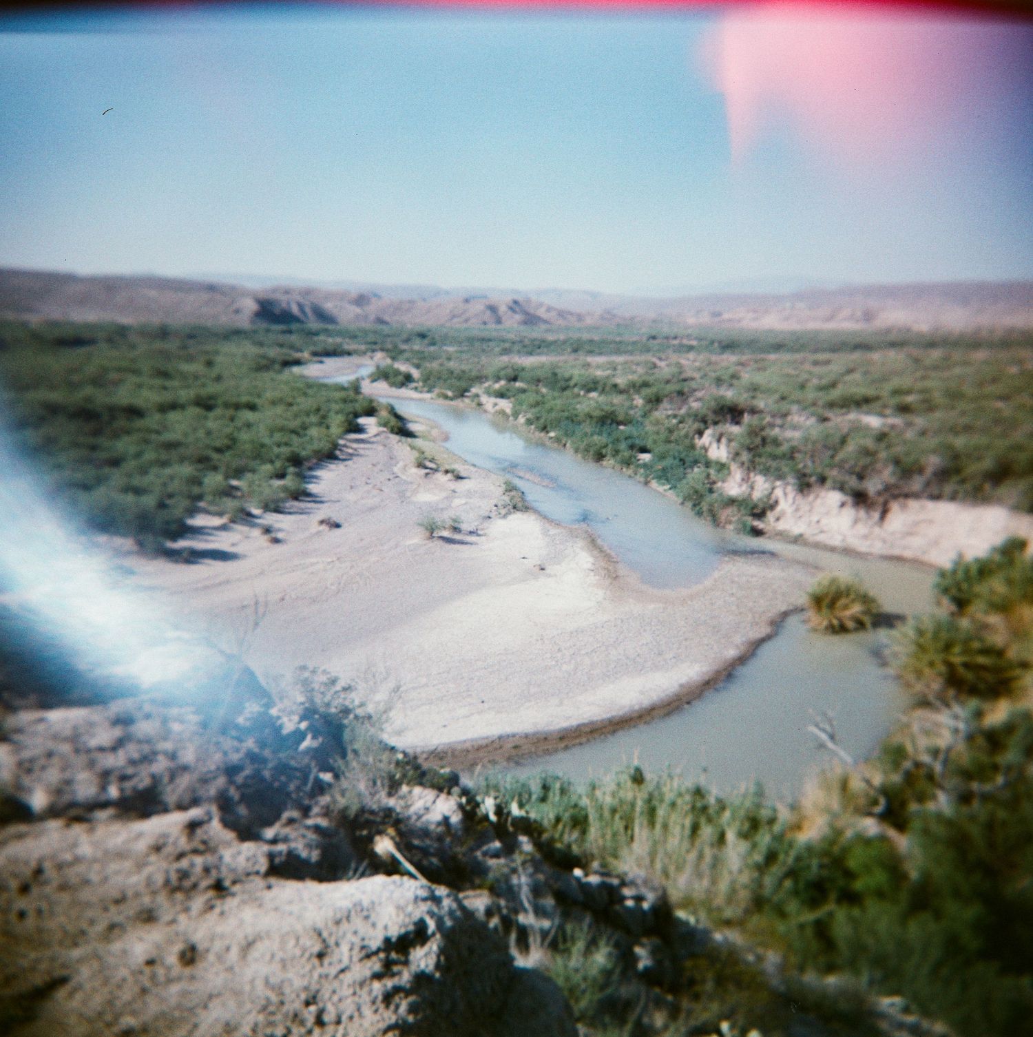 Aerial view of a winding river through arid desert landscape with green vegetation and sandy banks under a hazy sky.