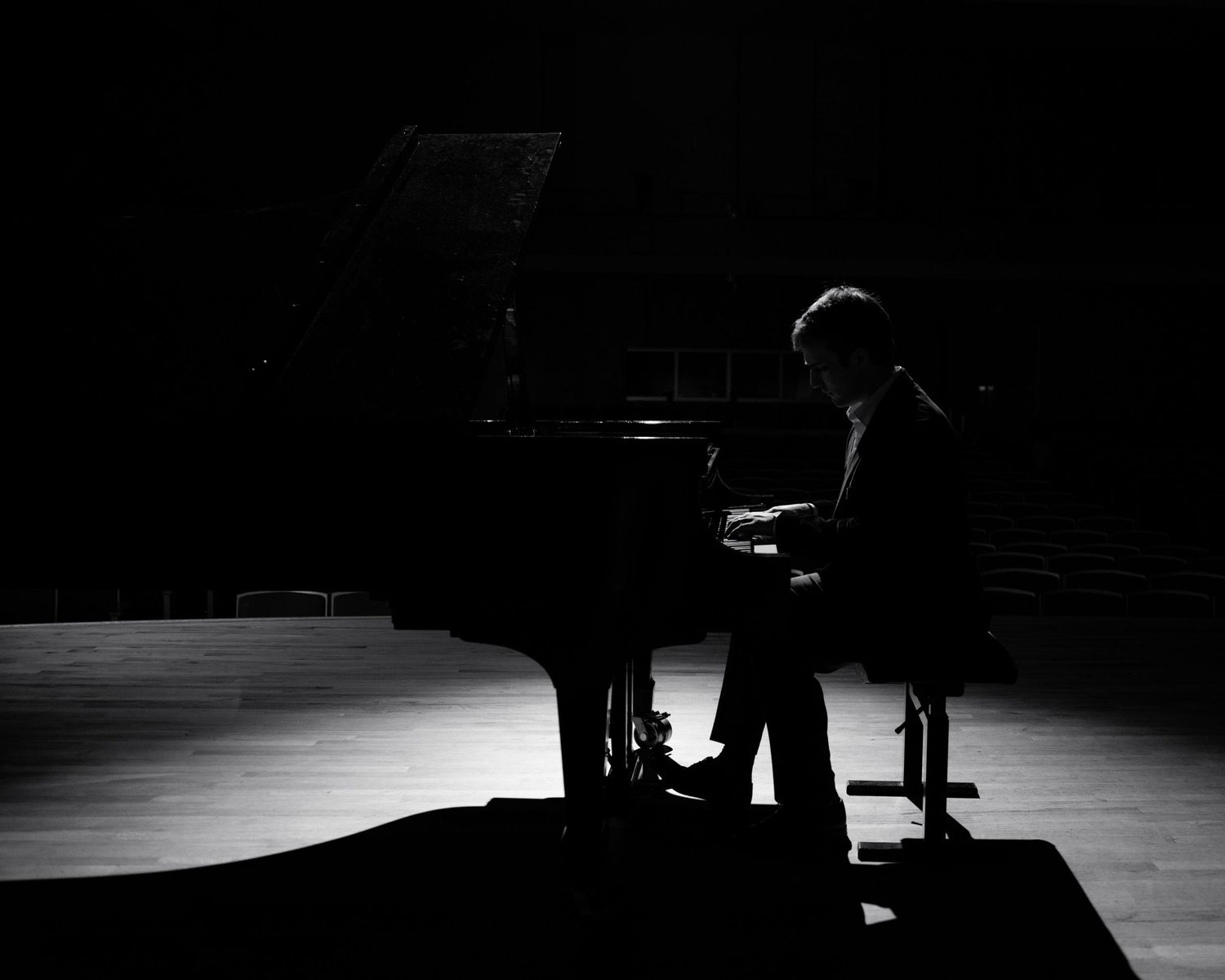 Silhouette of pianist performing solo on grand piano in darkened concert hall.