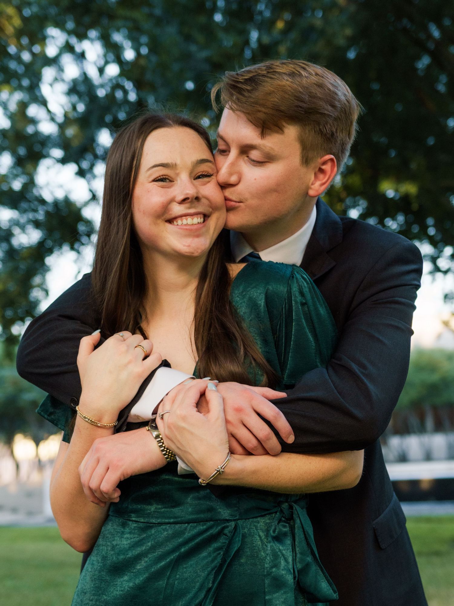 Man in dark suit kisses woman's cheek while embracing her from behind, woman in green velvet dress smiling.