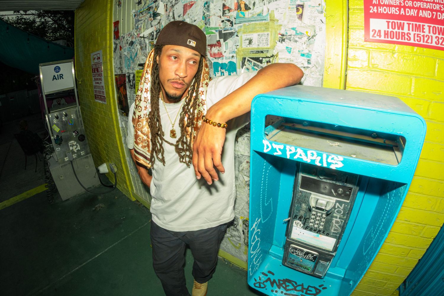 Man with dreadlocks in streetwear leans against a vintage blue payphone outside a vibrant, graffiti-accented urban convenience store.