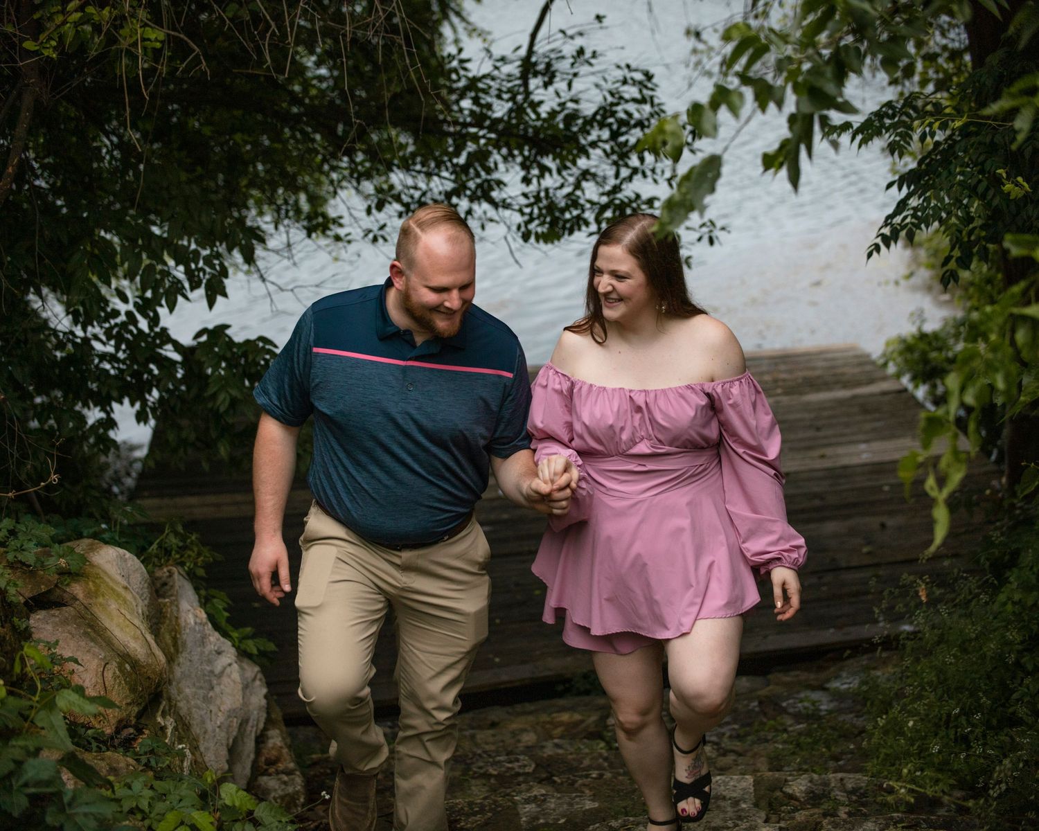 Couple walking hand in hand along wooded waterfront path during engagement photo session.